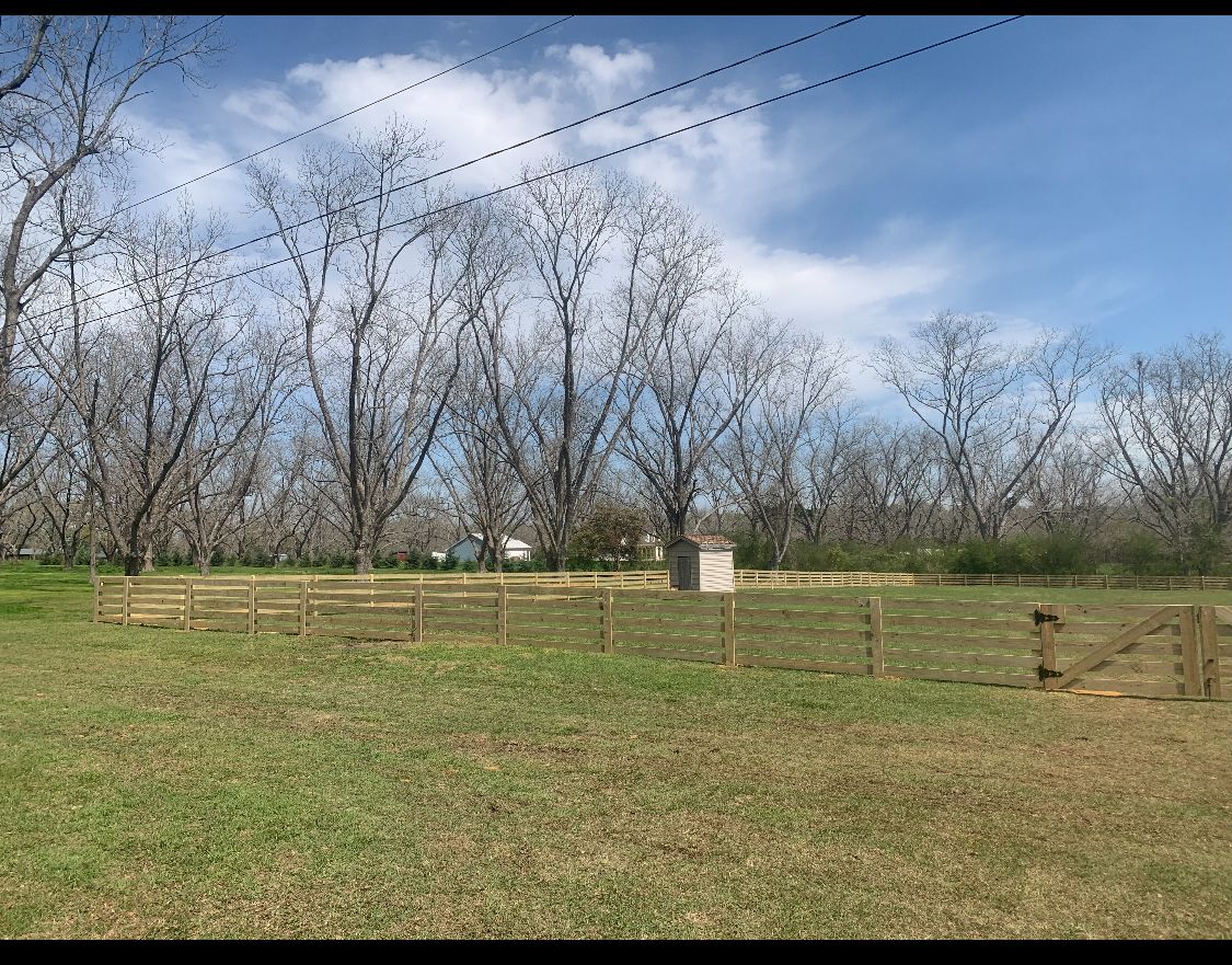 Wooden fence encloses a grassy area with trees and a small structure under a blue sky.