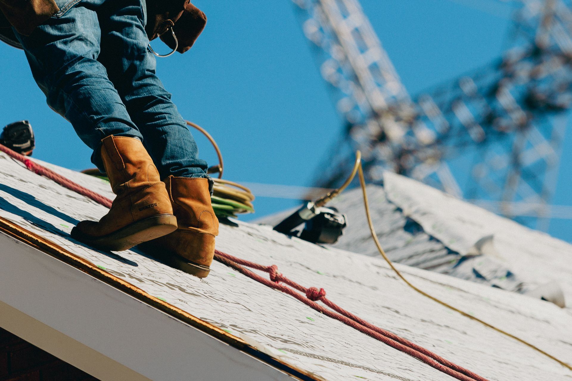 Construction worker on a rooftop, wearing boots and jeans, with safety ropes and a blue sky in the background.