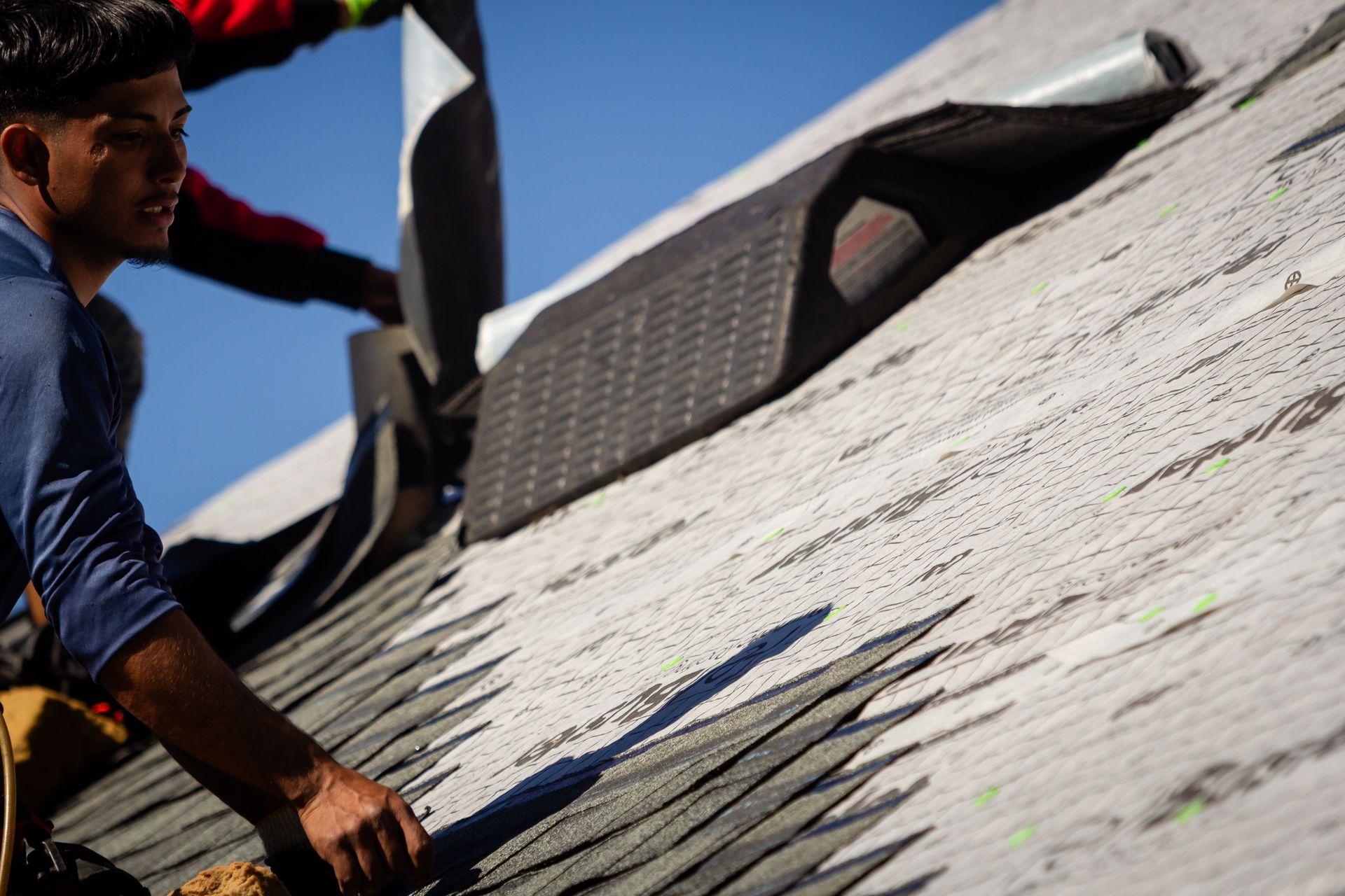 Man on rooftop installing roofing material. Blue sky, shingles, and tools are visible.