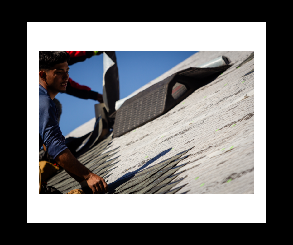 Workers repairing a roof; one holds up rolled black roofing material, blue sky in the background.
