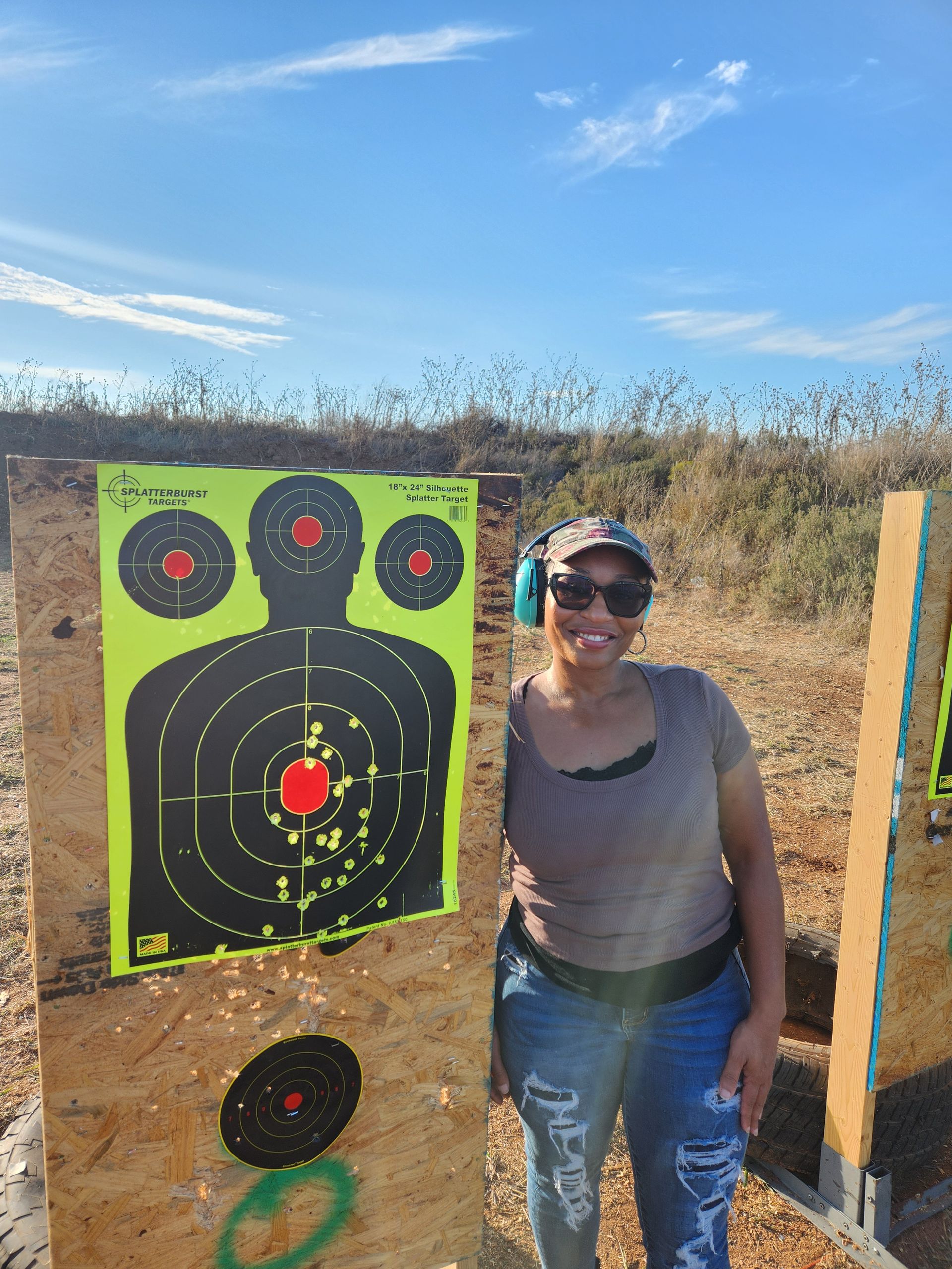 Woman at a shooting range smiles near a target with bullet holes. Blue sky, outdoor setting.