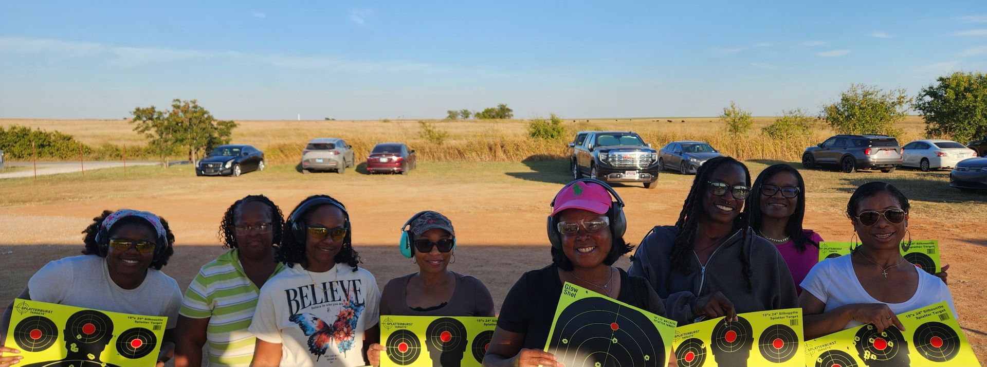 Group of women holding up shooting targets at an outdoor range, smiles present, cars in the background.