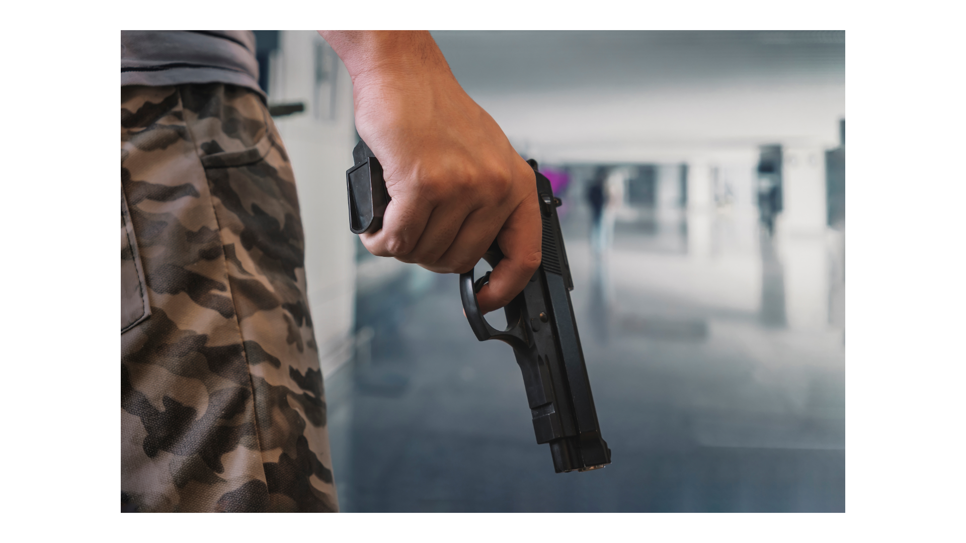Close-up of a hand holding a handgun beside camouflage pants in a bright indoor hallway.