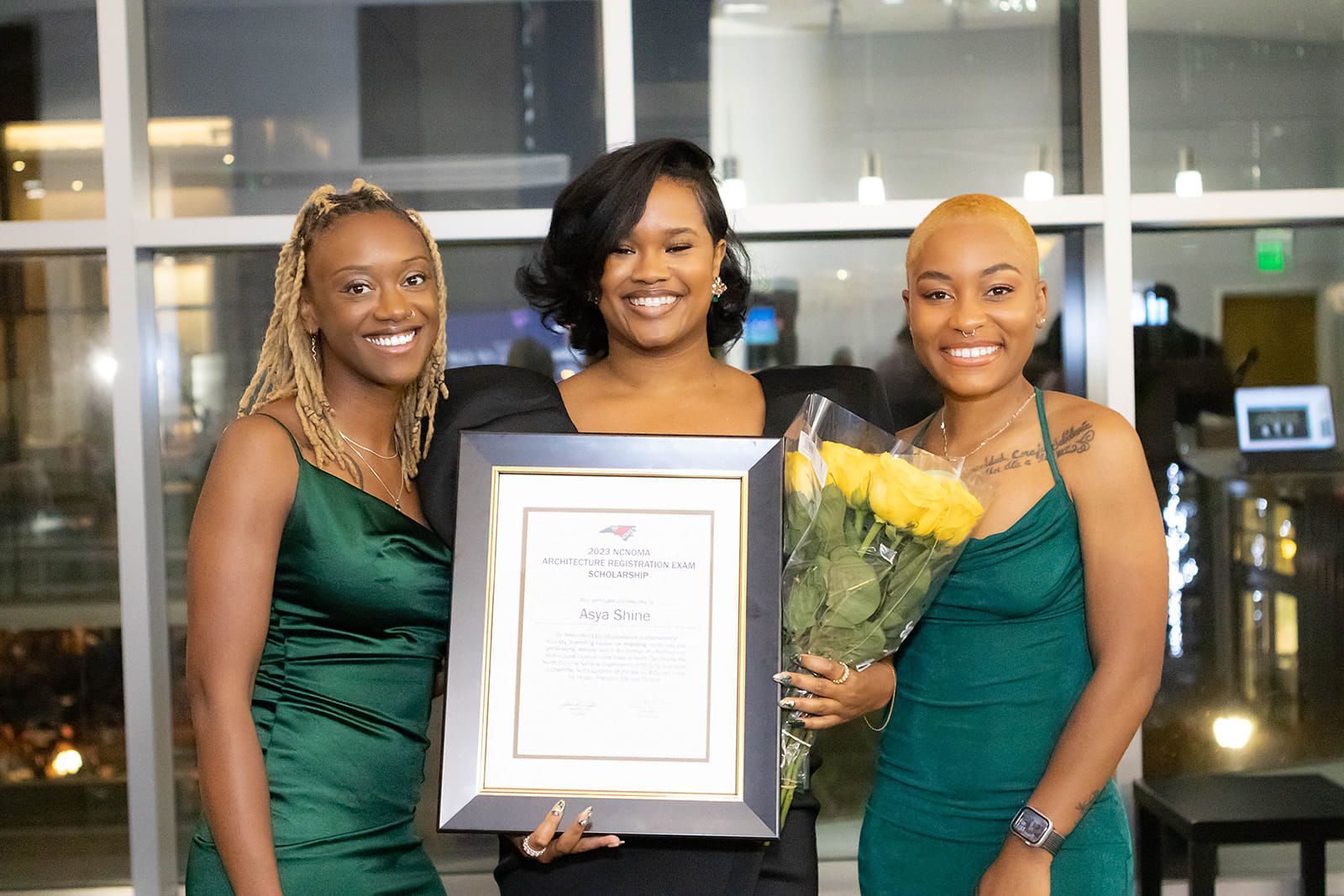 Three women in green dresses are standing next to each other holding a certificate and flowers.