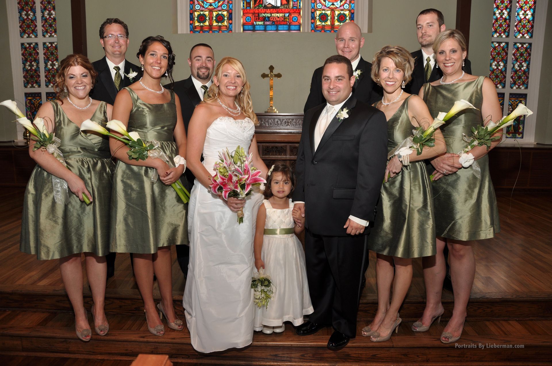 A bride and groom pose with their wedding party in front of a stained glass window