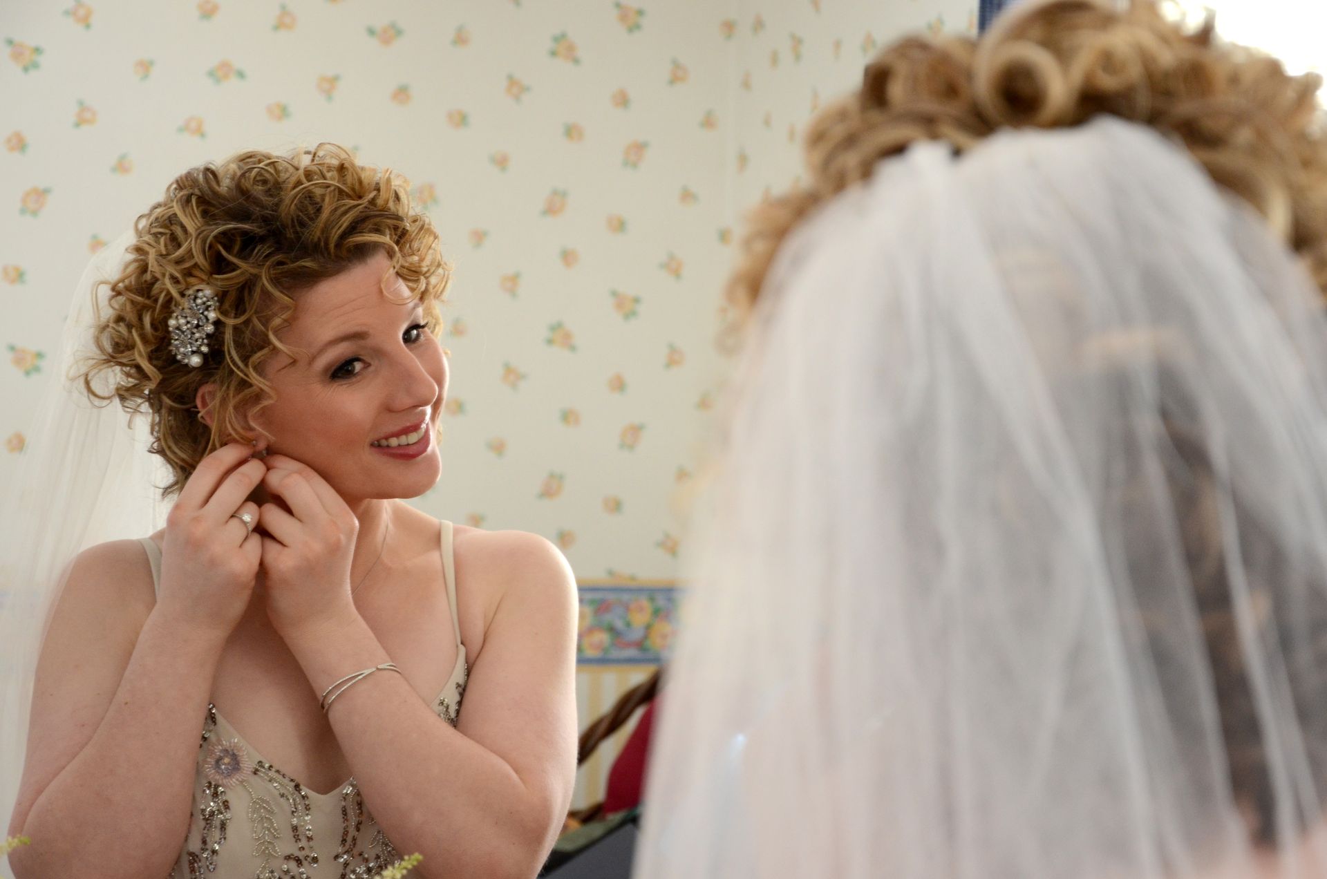 A woman in a wedding dress is putting on her earrings in front of a mirror.