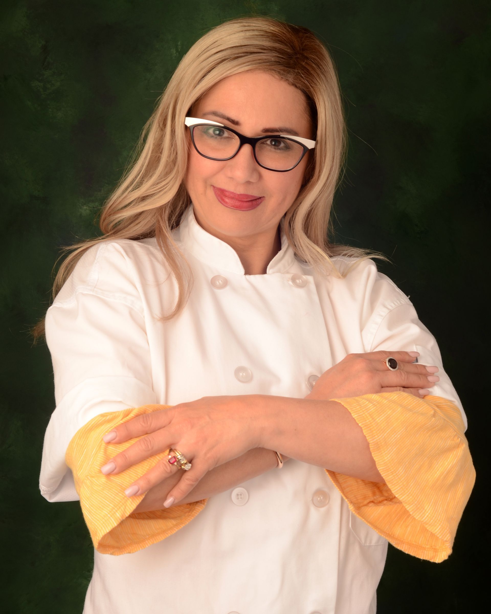 A woman wearing glasses and a chef 's coat is standing with her arms crossed.
