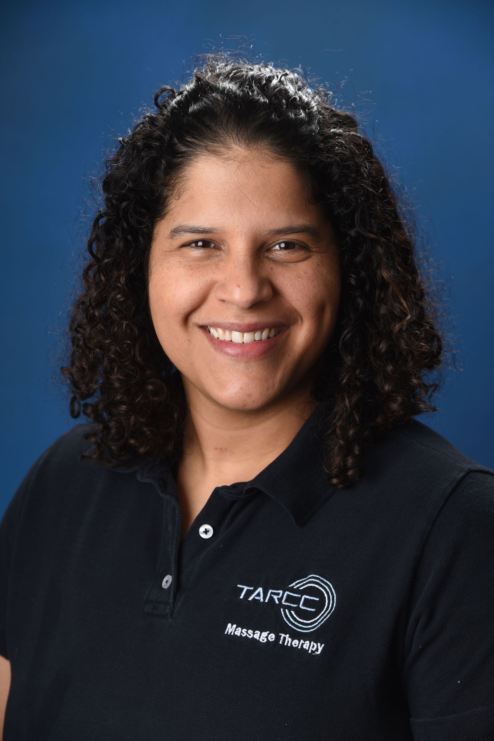 A woman with curly hair is wearing a black polo shirt and smiling.