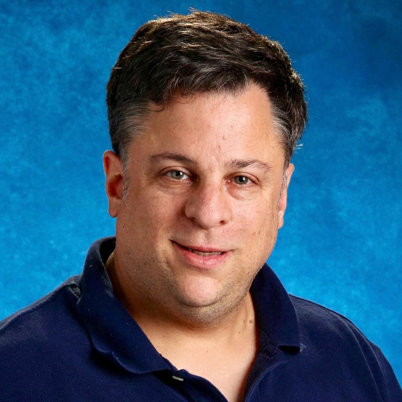 A man in a blue shirt is smiling in front of a blue background