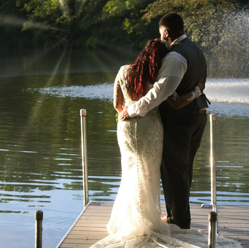 A bride and groom are hugging on a dock overlooking a lake