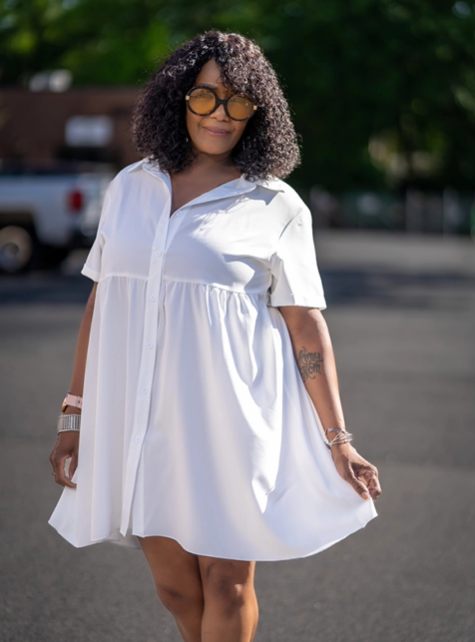 A woman wearing a white dress and sunglasses is standing in a parking lot.