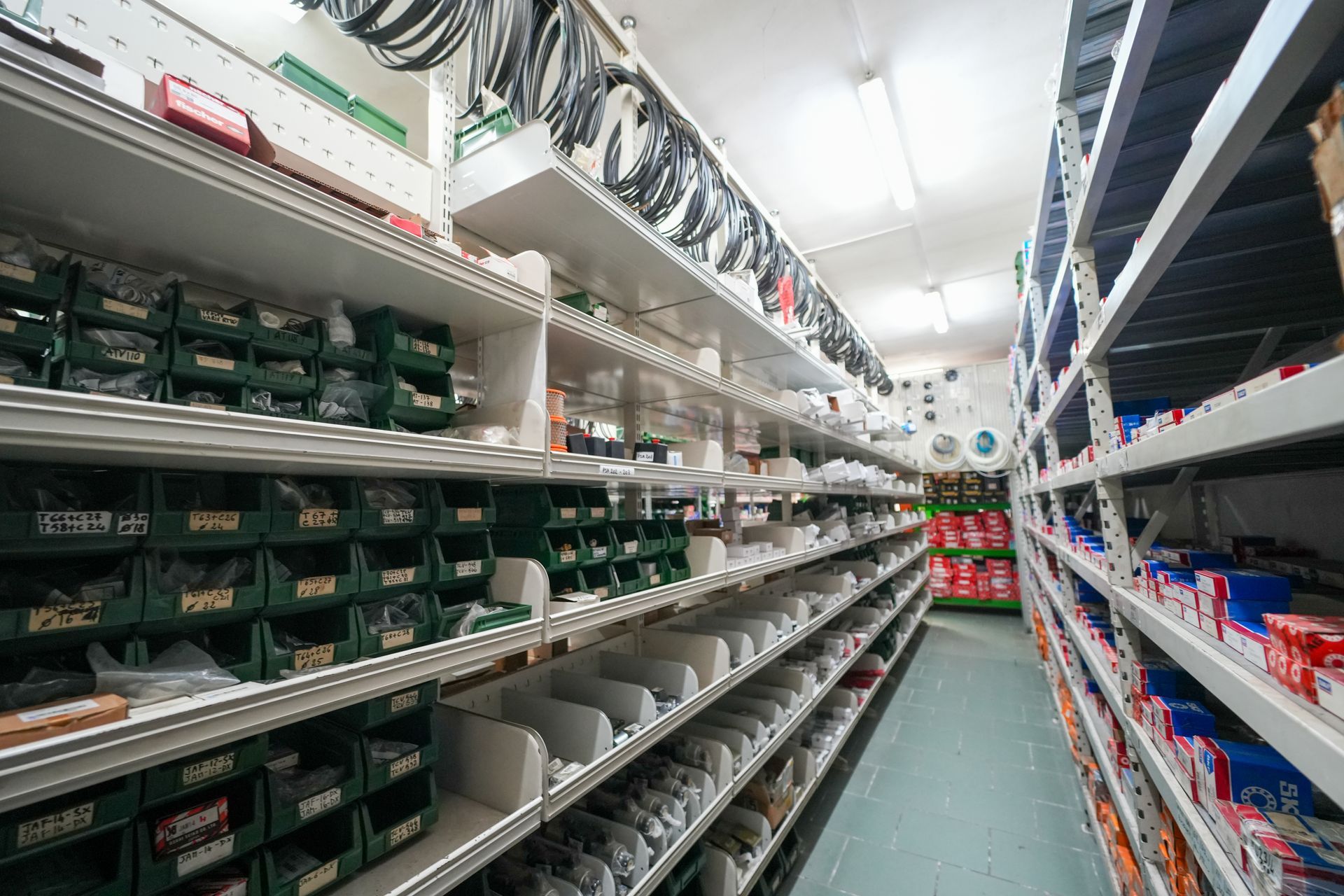 Uno stretto corridoio in un magazzino industriale, fiancheggiato da scaffalature metalliche piene di piccoli componenti in contenitori e scatole verdi.