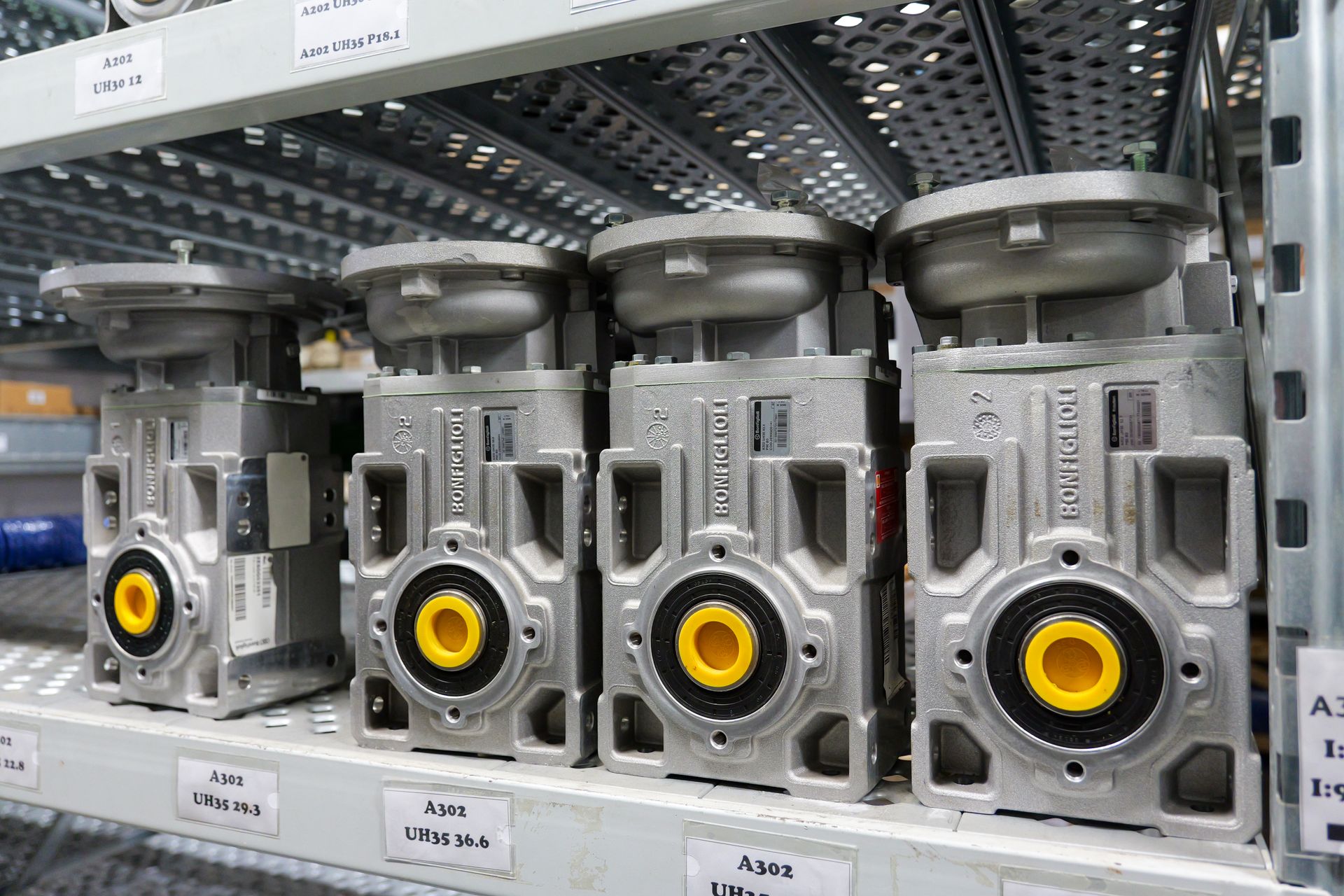 Quattro motoriduttori industriali identici, con involucri in metallo argentato e cappucci gialli, sono posizionati su una scaffalatura metallica.