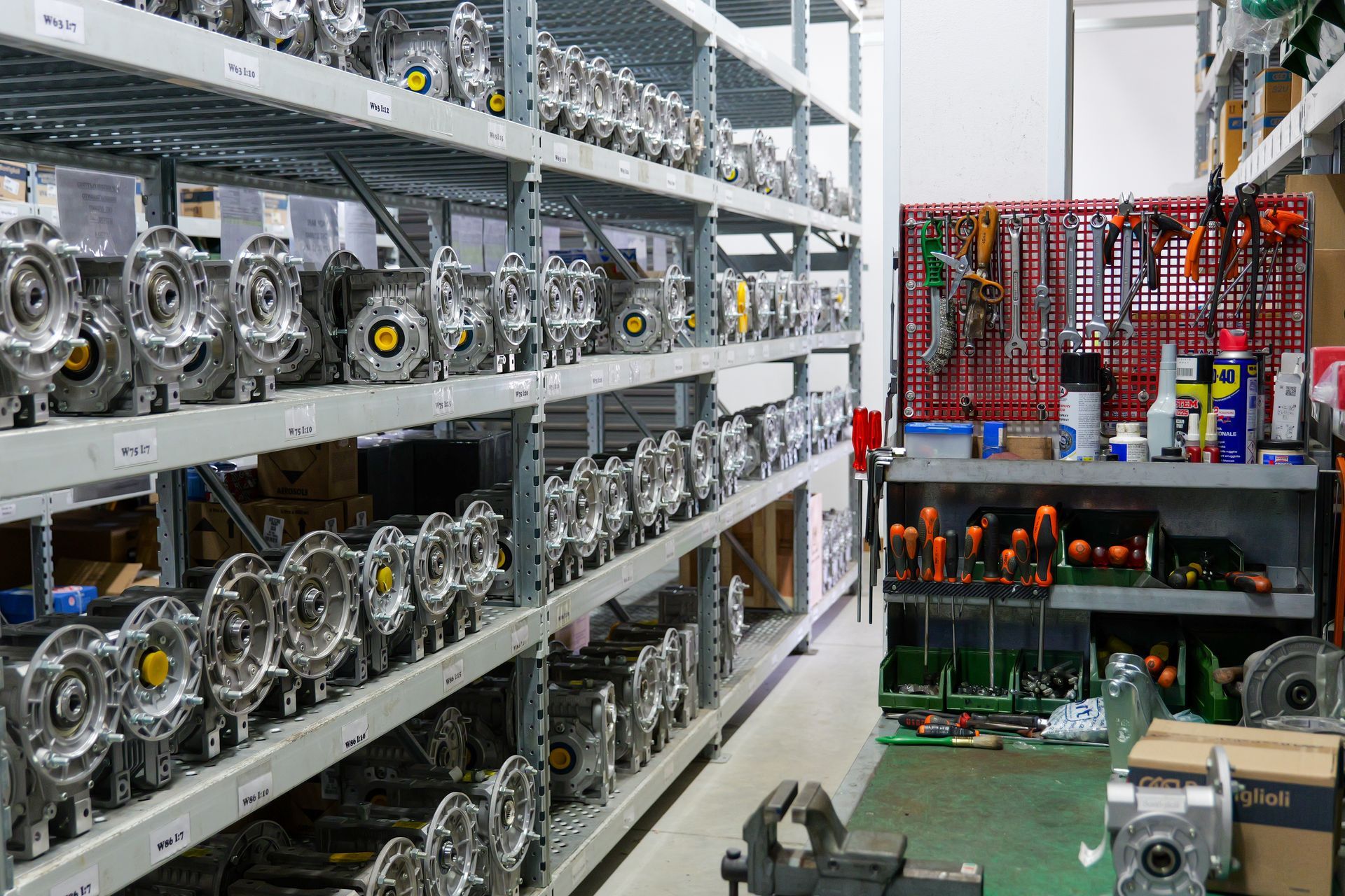 Scaffalature piene di componenti meccanici industriali accanto a una postazione di lavoro con una parete attrezzata rossa e vari utensili.