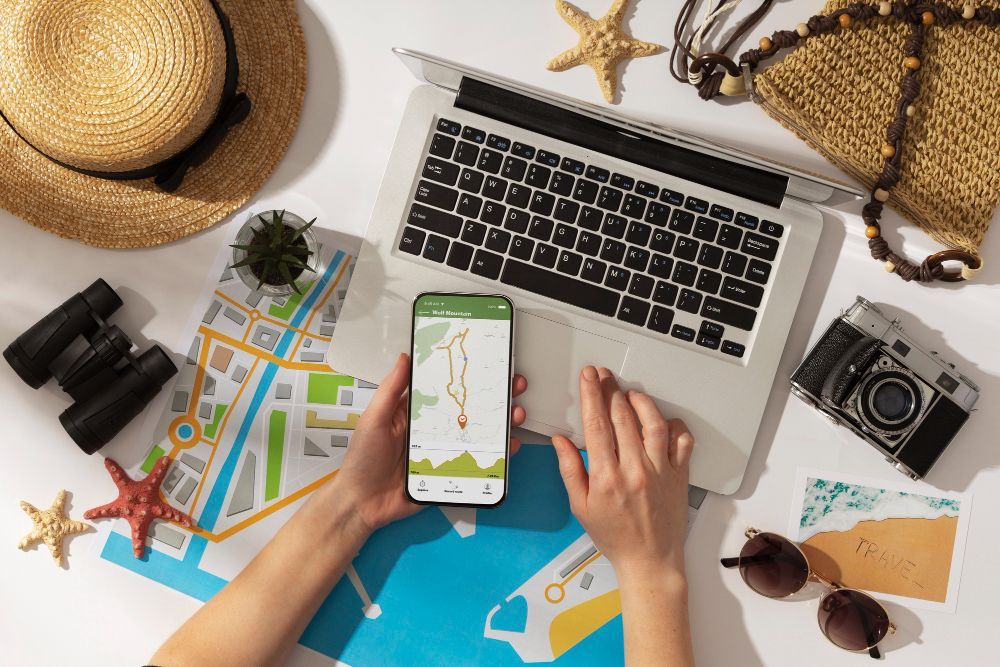 Hands using a smartphone with a map app on a laptop surrounded by travel items: hat, binoculars, camera, sunglasses.
