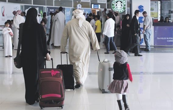 People at an airport, some wearing traditional Arab clothing, walking with luggage.