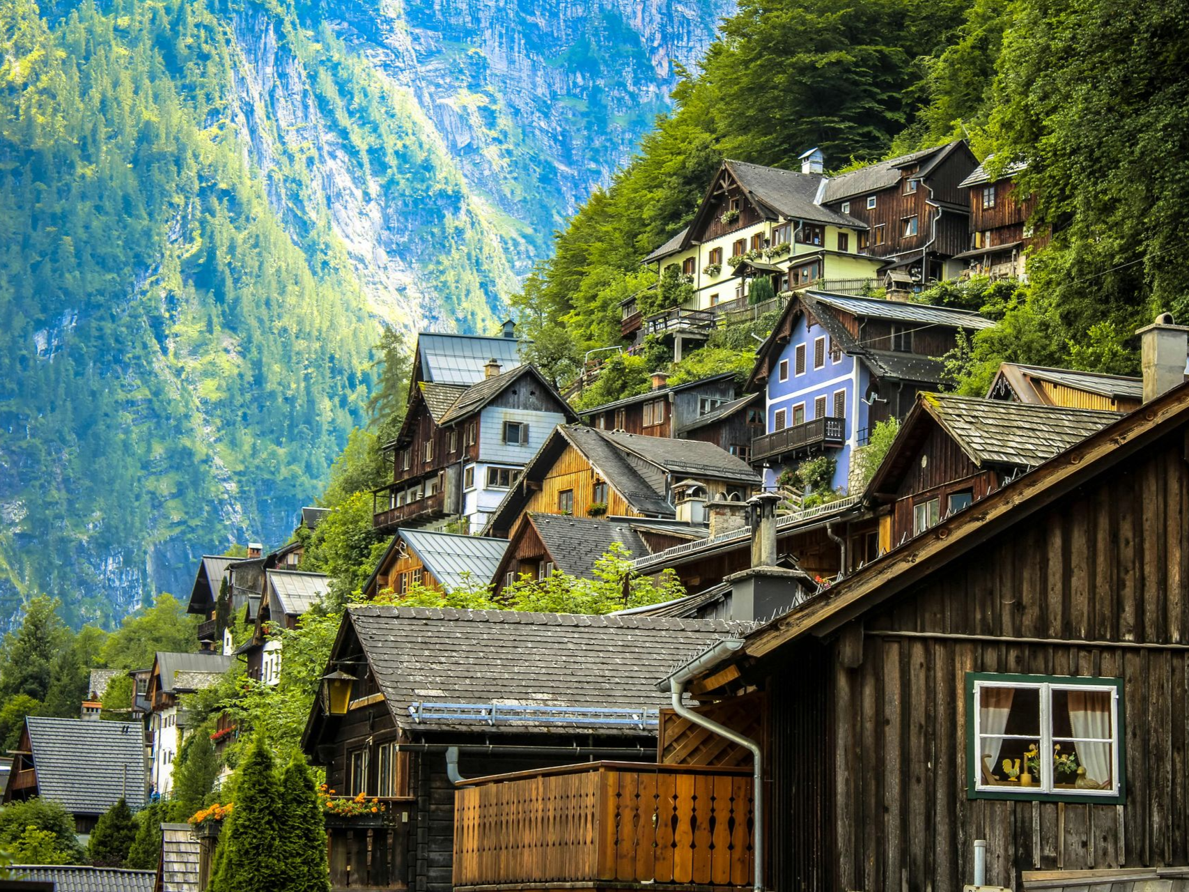 Wooden houses nestled on a mountain slope in Hallstatt, Austria, with a backdrop of green trees and a blue sky.