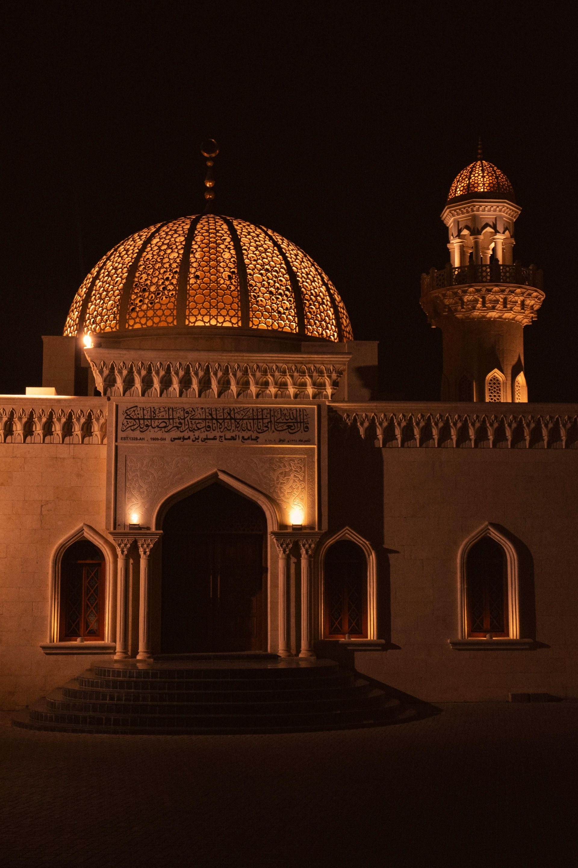 Mosque illuminated at night, dome and minaret aglow, arched entrance, warm colors, stone structure.