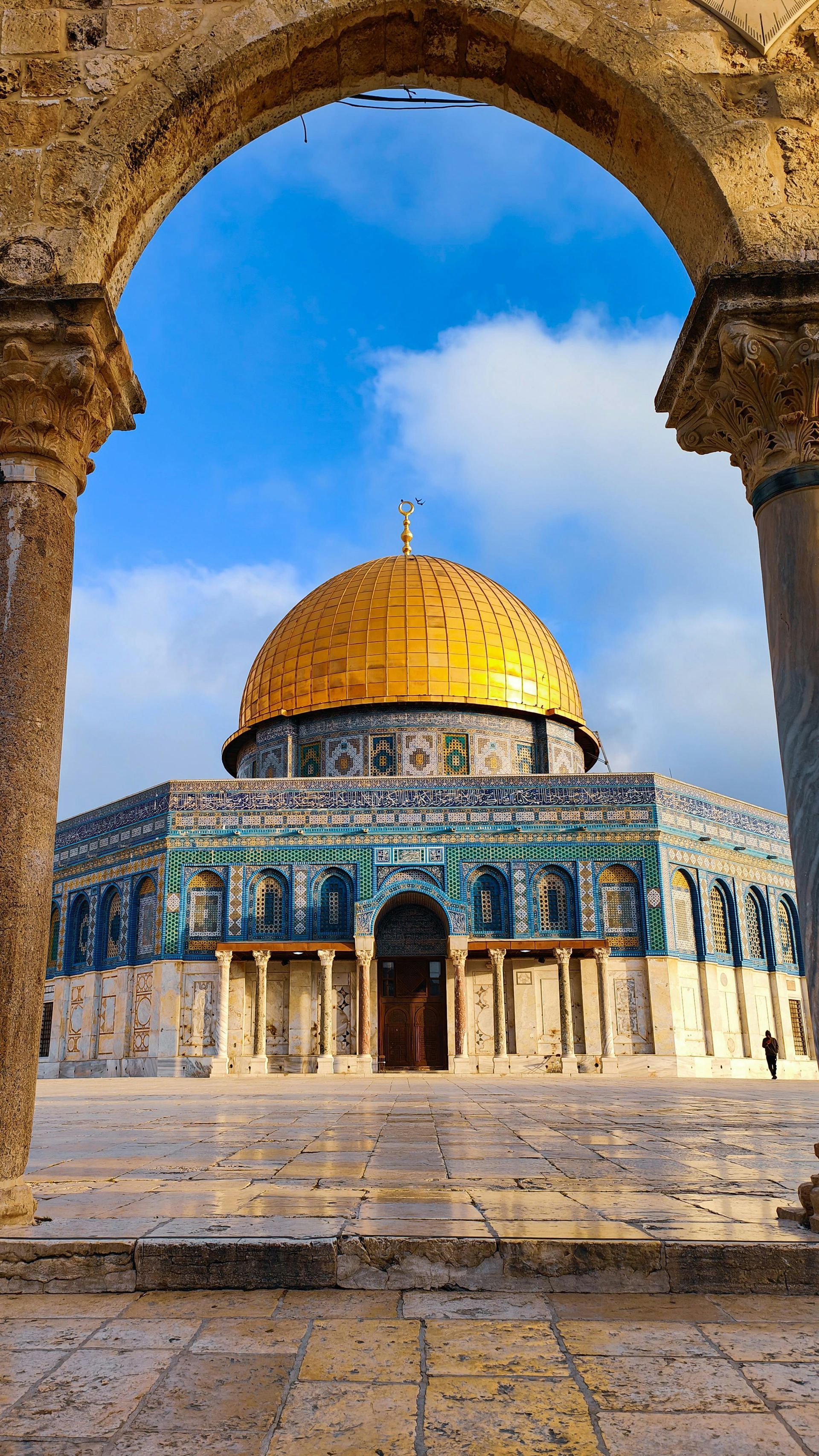 Dome of the Rock, Jerusalem, framed by an archway. Gold dome, blue and white tiled walls, against a blue sky.