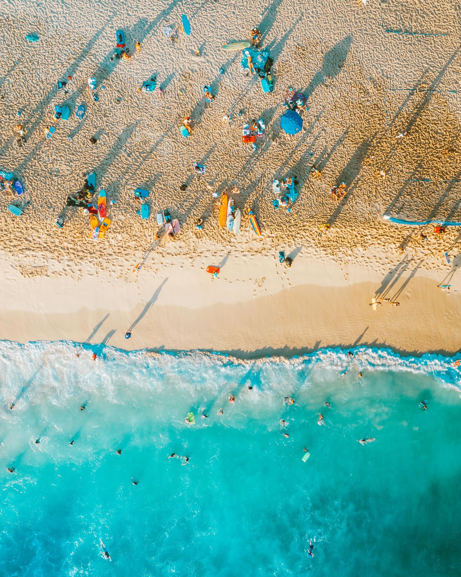 Aerial view of beach with people swimming in turquoise water, sunbathing on sand.