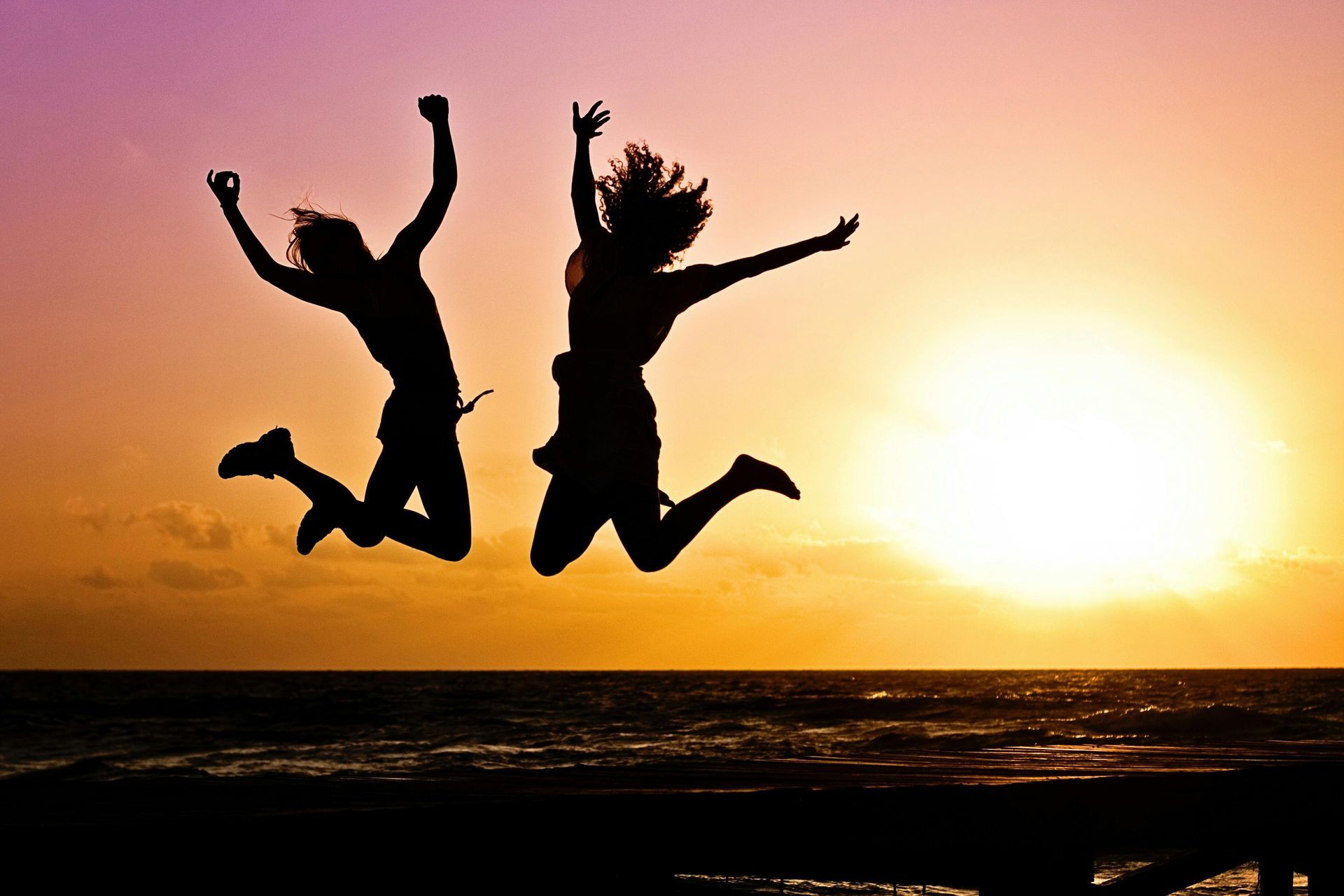 Two silhouetted people jumping with arms raised against a colorful sunset over water.