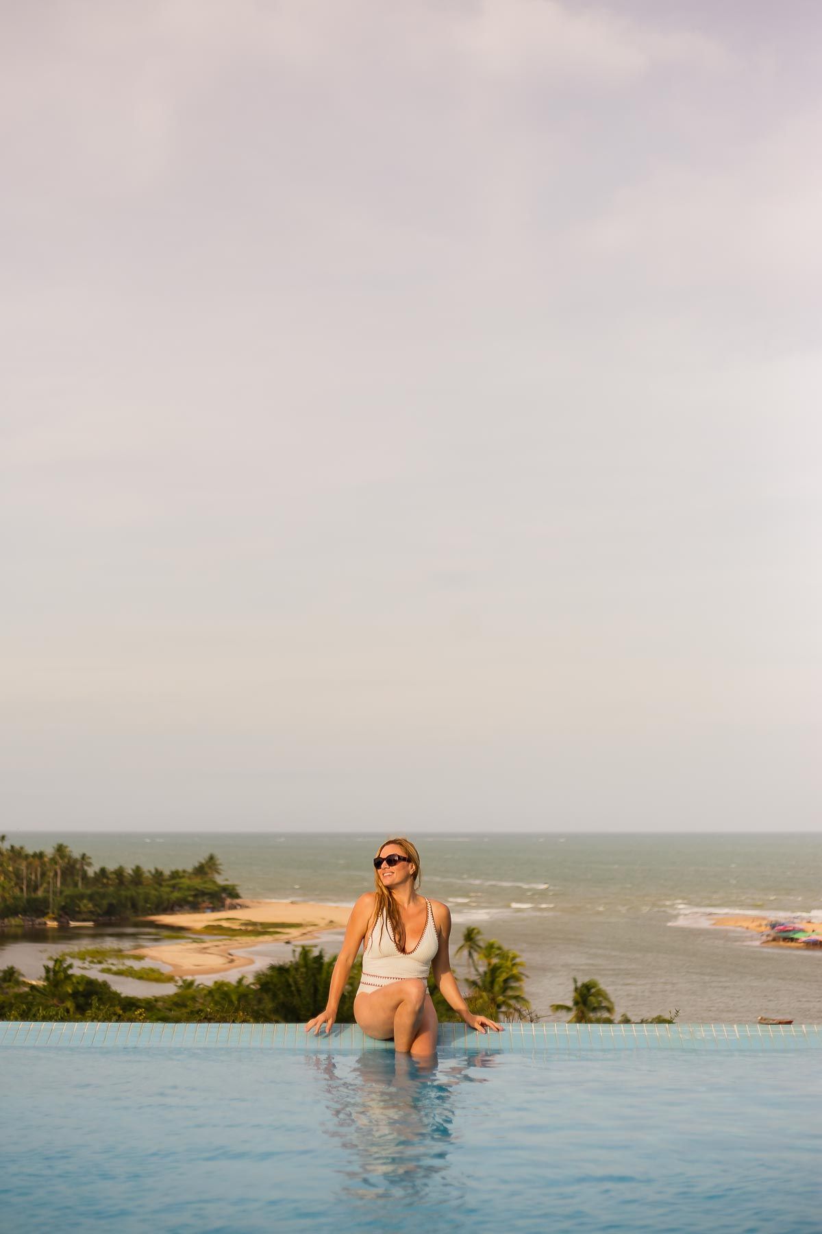 Flores rosas brilhantes emolduram uma piscina turquesa em um deck de madeira; árvores e céu azul ao fundo.