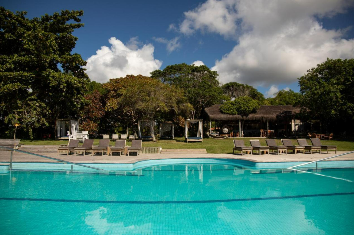 Piscina com espreguiçadeiras, árvores e um prédio sob um céu azul com nuvens brancas e fofas.