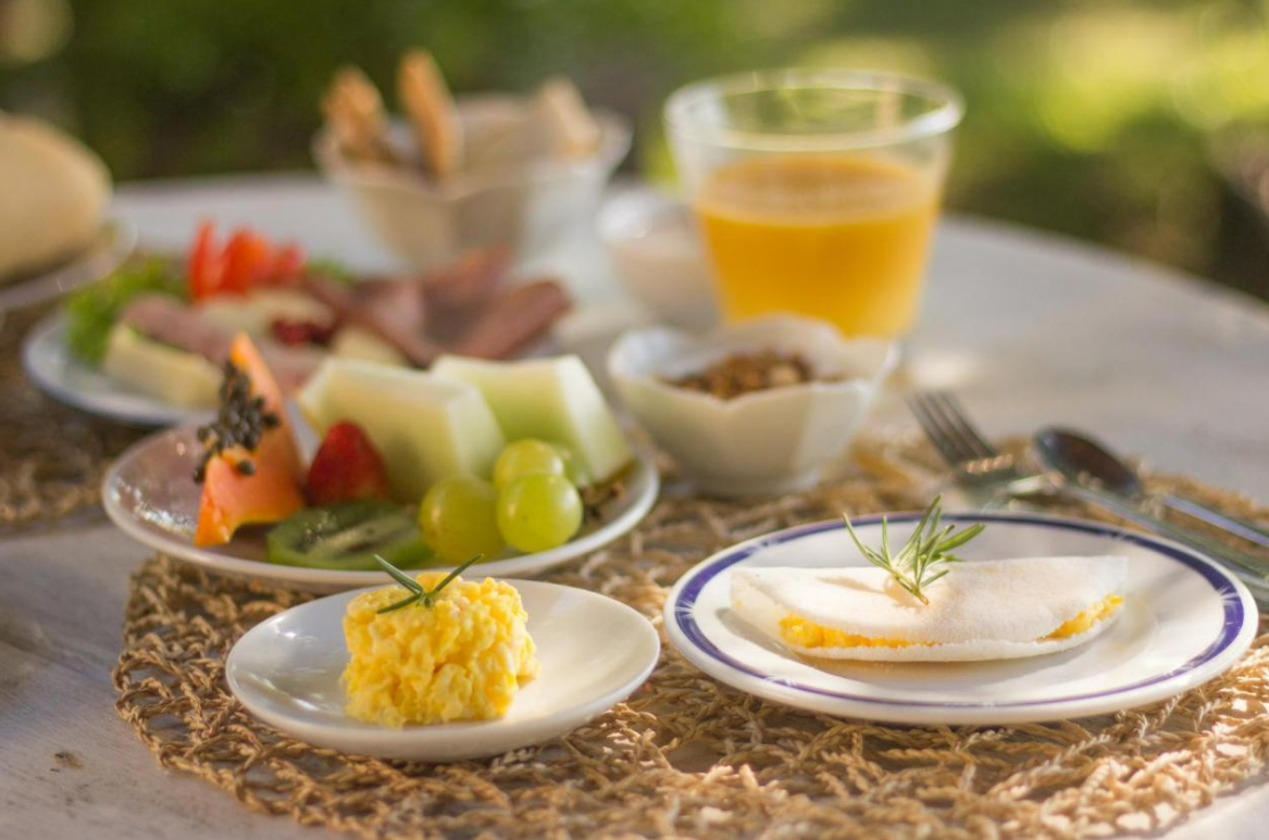 Café da manhã servido ao ar livre: frutas, carnes, queijos, sucos, pães, doces e ovos mexidos salgados.