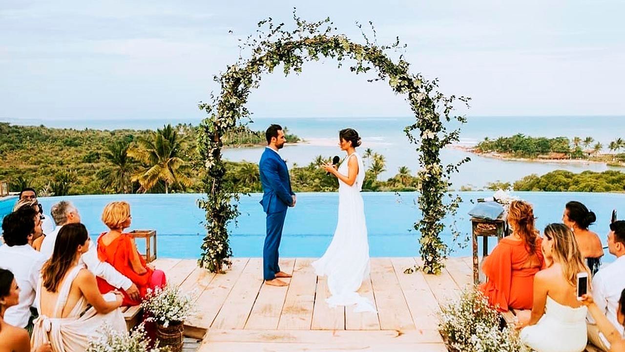 Cerimônia de casamento à beira de uma piscina com vista para o mar. A noiva lê os votos sob um arco floral. Os convidados assistem.