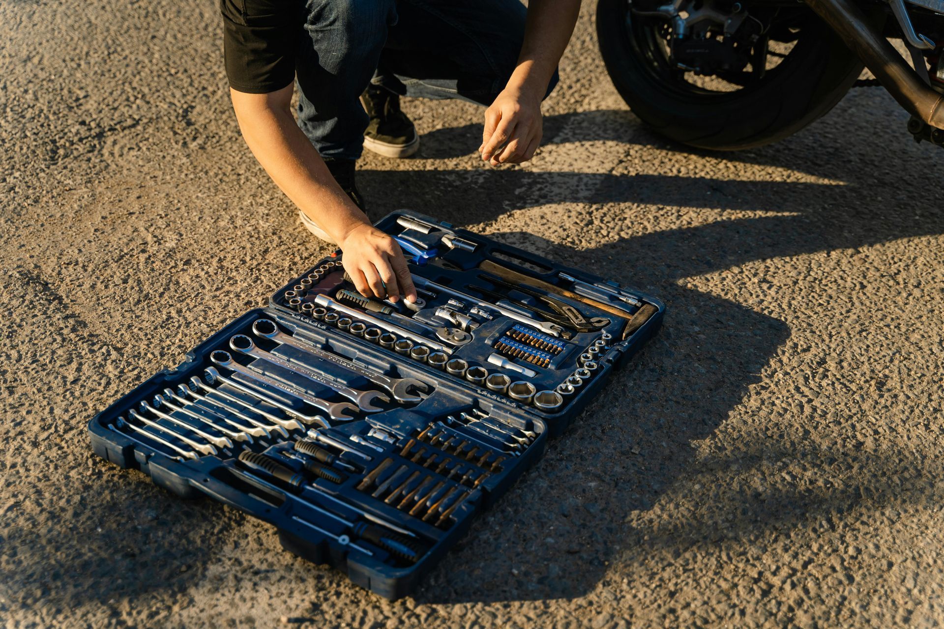 Person kneeling next to a motorcycle, opening a blue toolbox on asphalt.