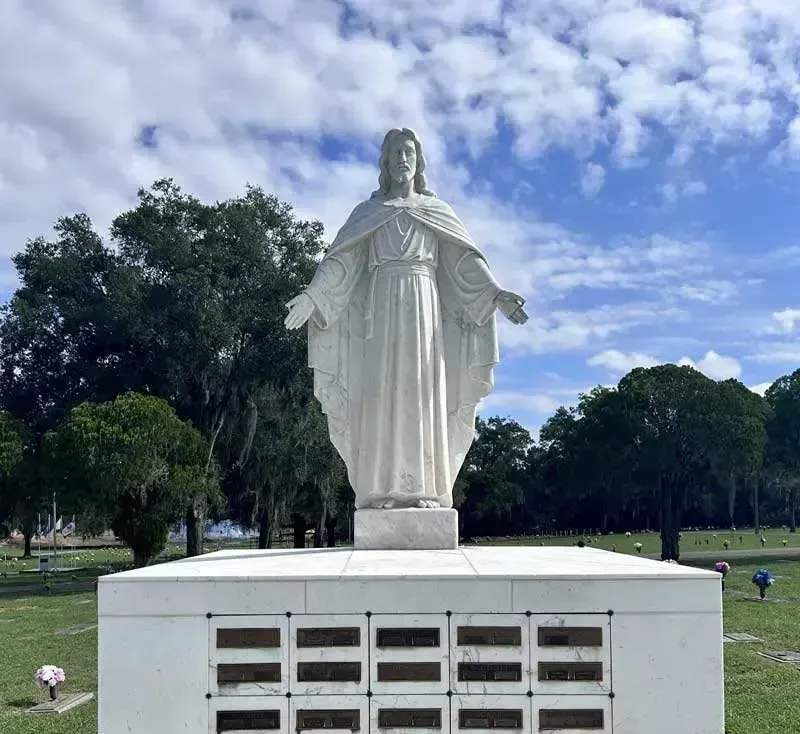 White statue of Jesus with open arms atop a white mausoleum in a cemetery, with trees and a cloudy sky.