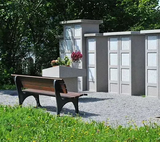 A bench sits in front of a gray mausoleum. Lush greenery and water in the background. Sunny day.