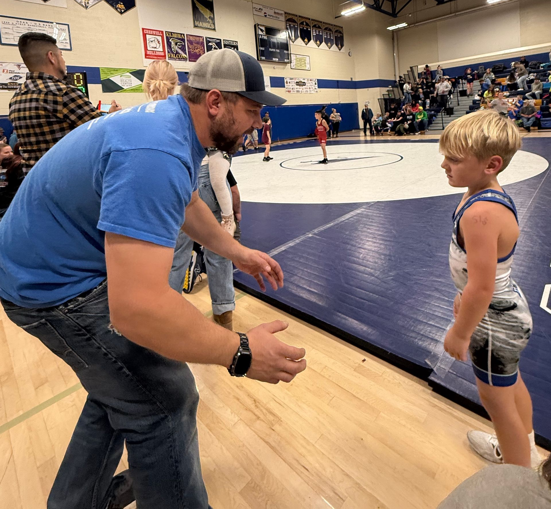 Coach crouching beside a young wrestler near a blue mat in a gymnasium during a match
