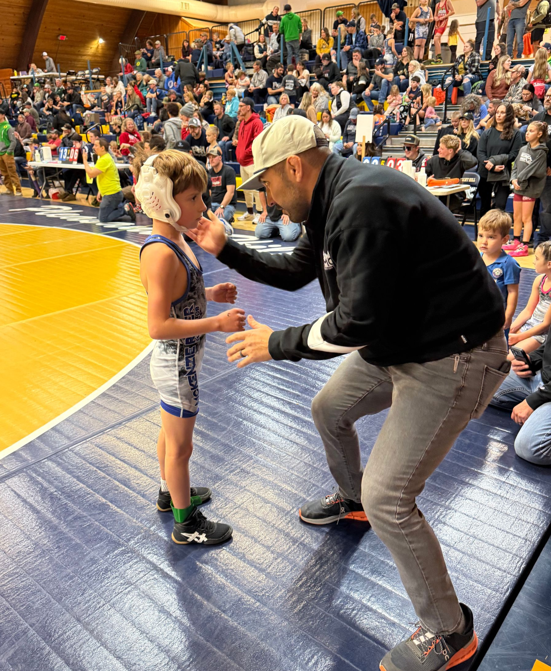 Coach kneels on wrestling mat, speaking to a young athlete, with a crowded gym bleacher in the background