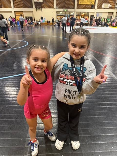 Two children in a gym posing on a wrestling mat with medals around their neck, smiling and holding up peace signs.