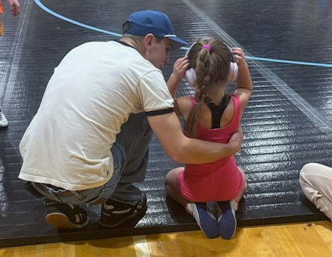 Man kneels beside a child on a wrestling mat, helping adjust her stance during practice