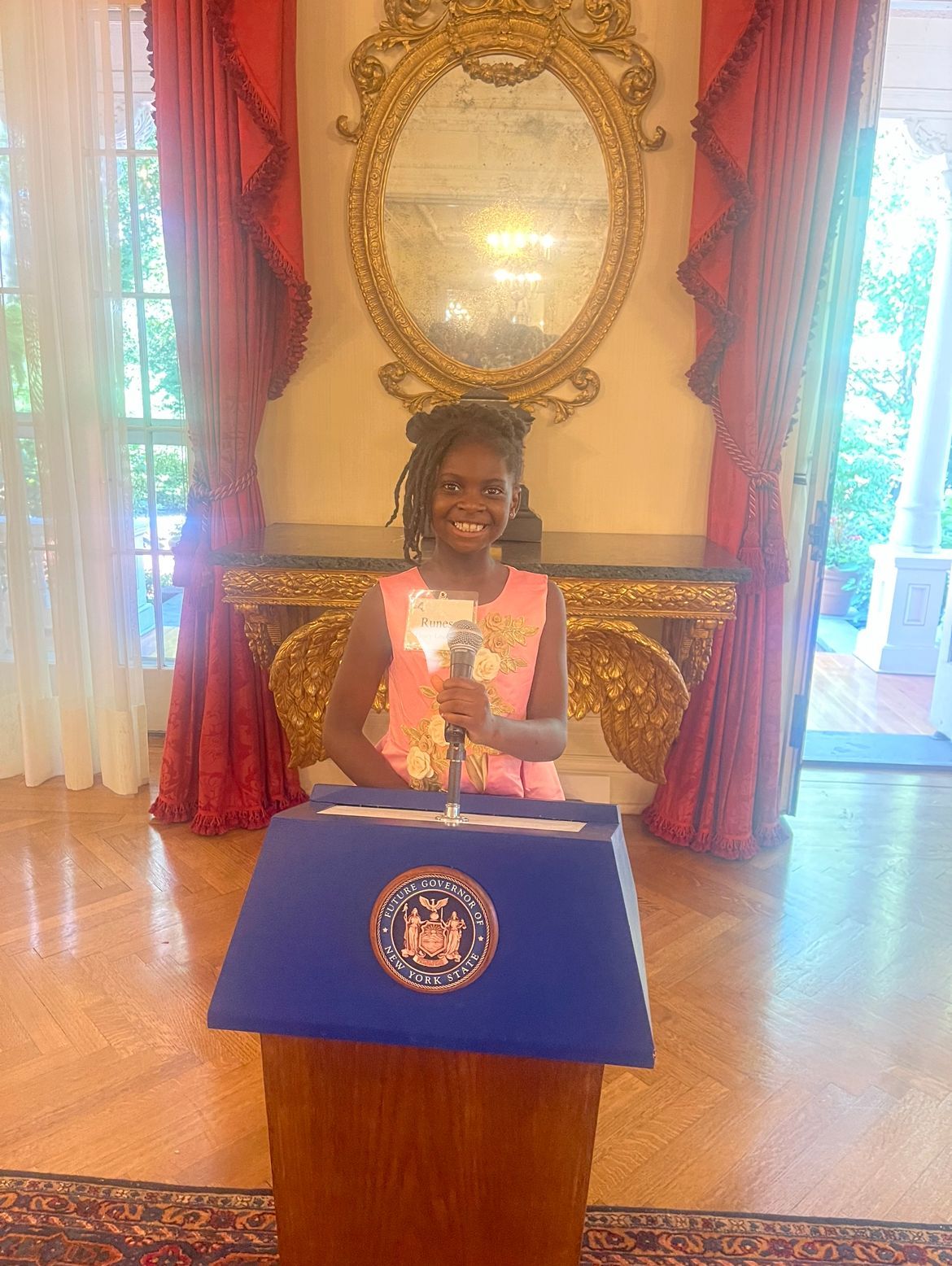 Young person speaking at a podium with a microphone, indoors near windows.