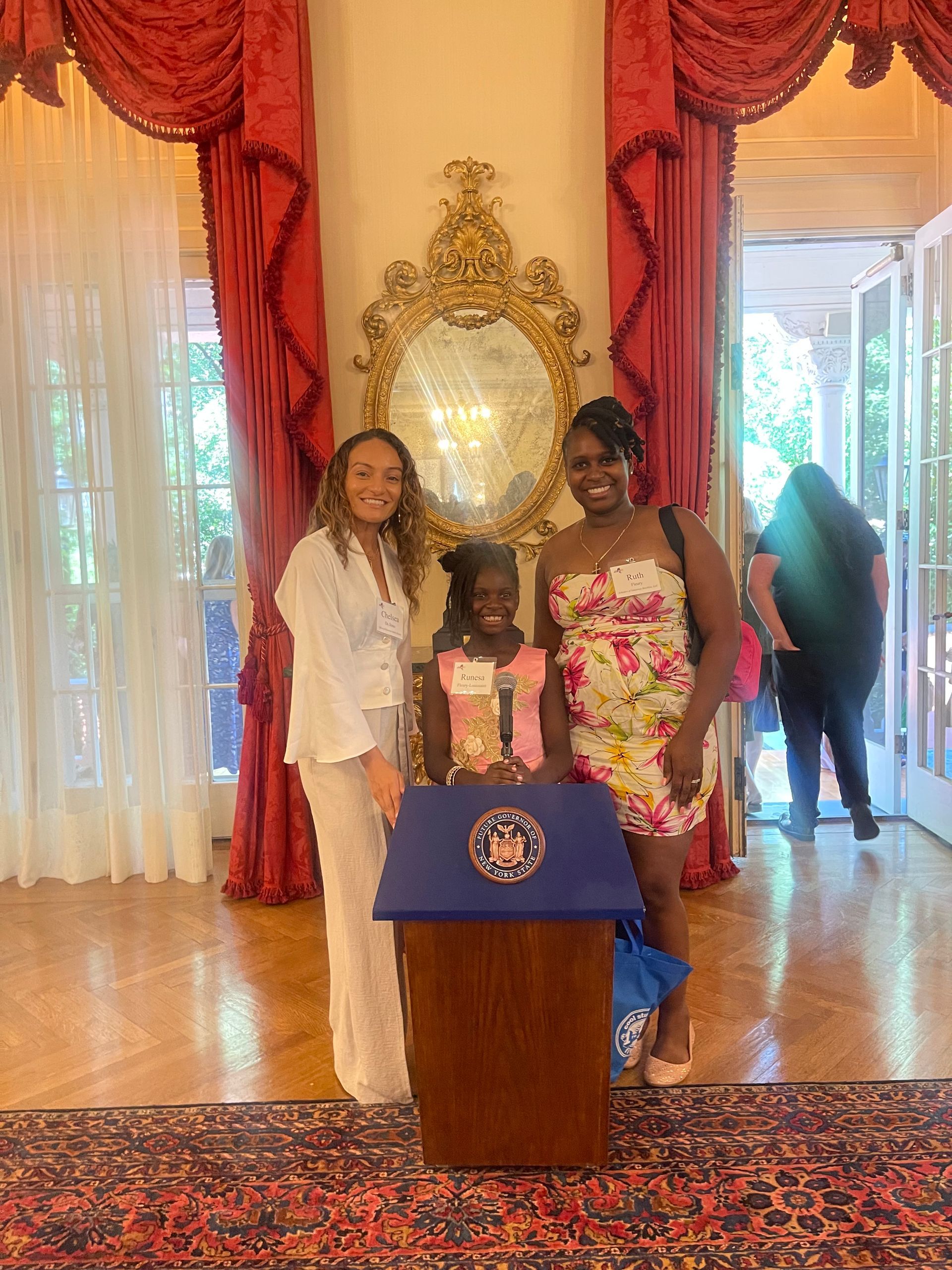 Two women and a child stand by a podium in a room with ornate decor.