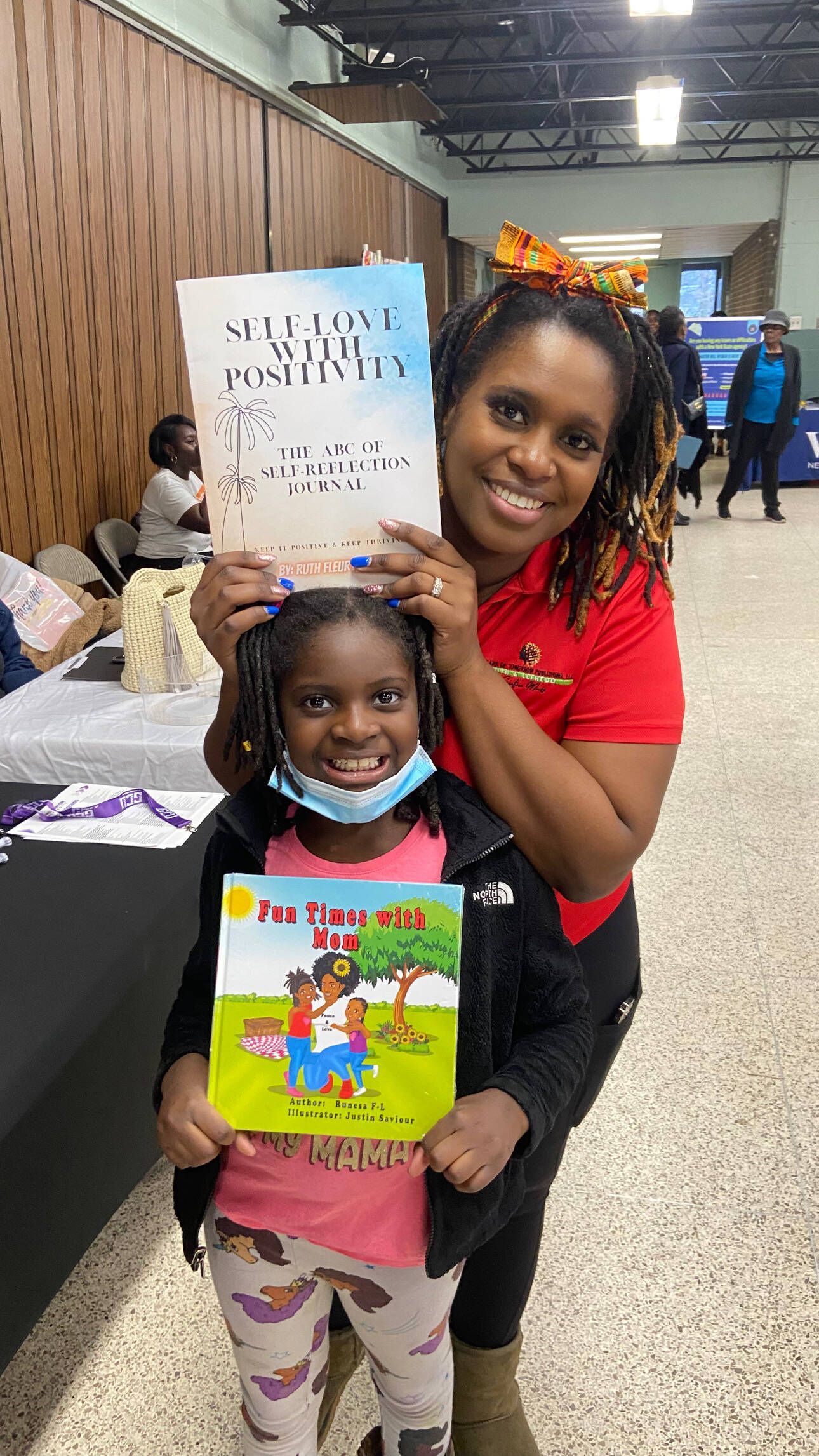 Woman and child holding books, smiling at the camera. Event setting with tables and other people.