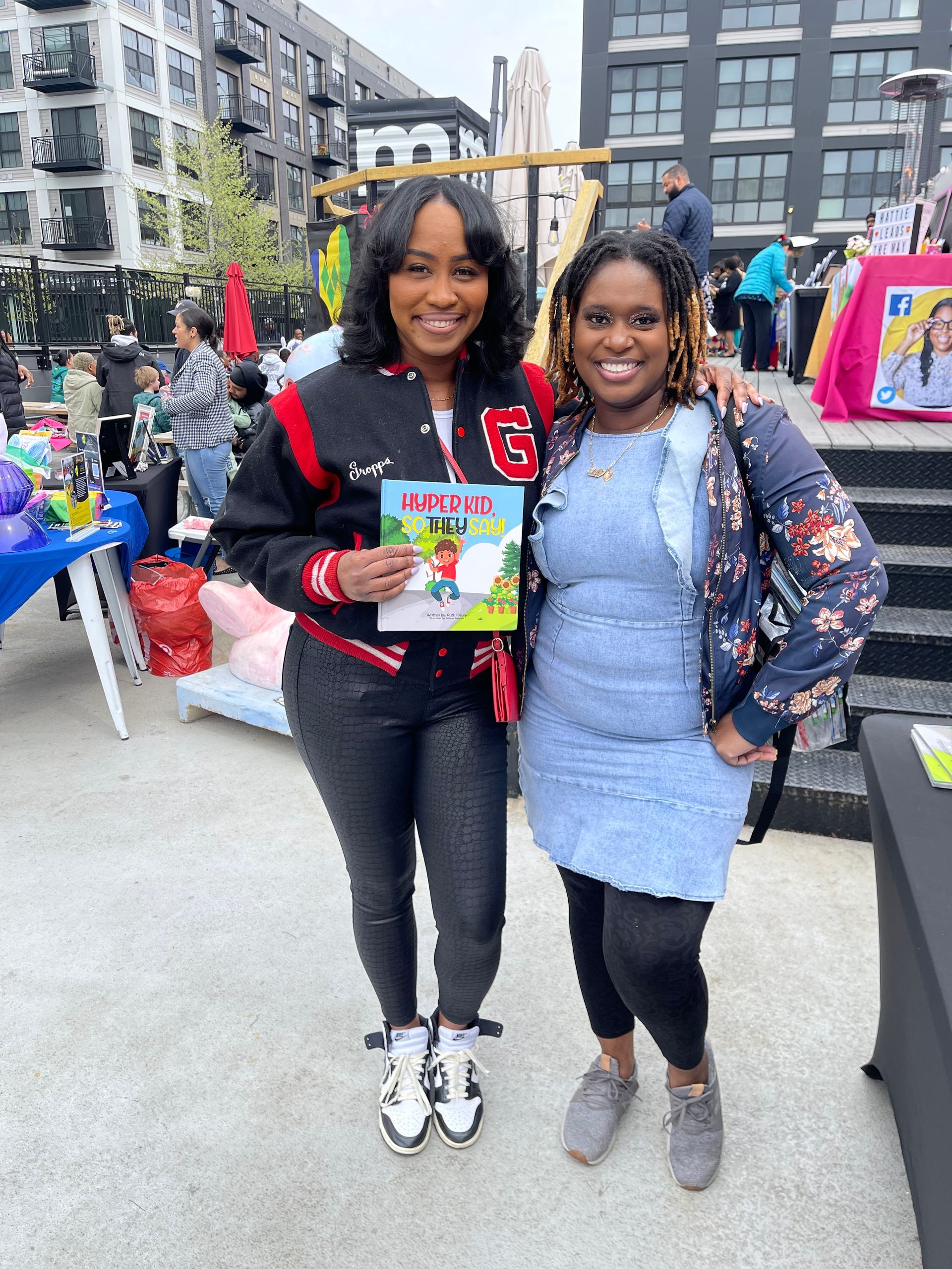 Two women smiling, holding a book outdoors at an event.