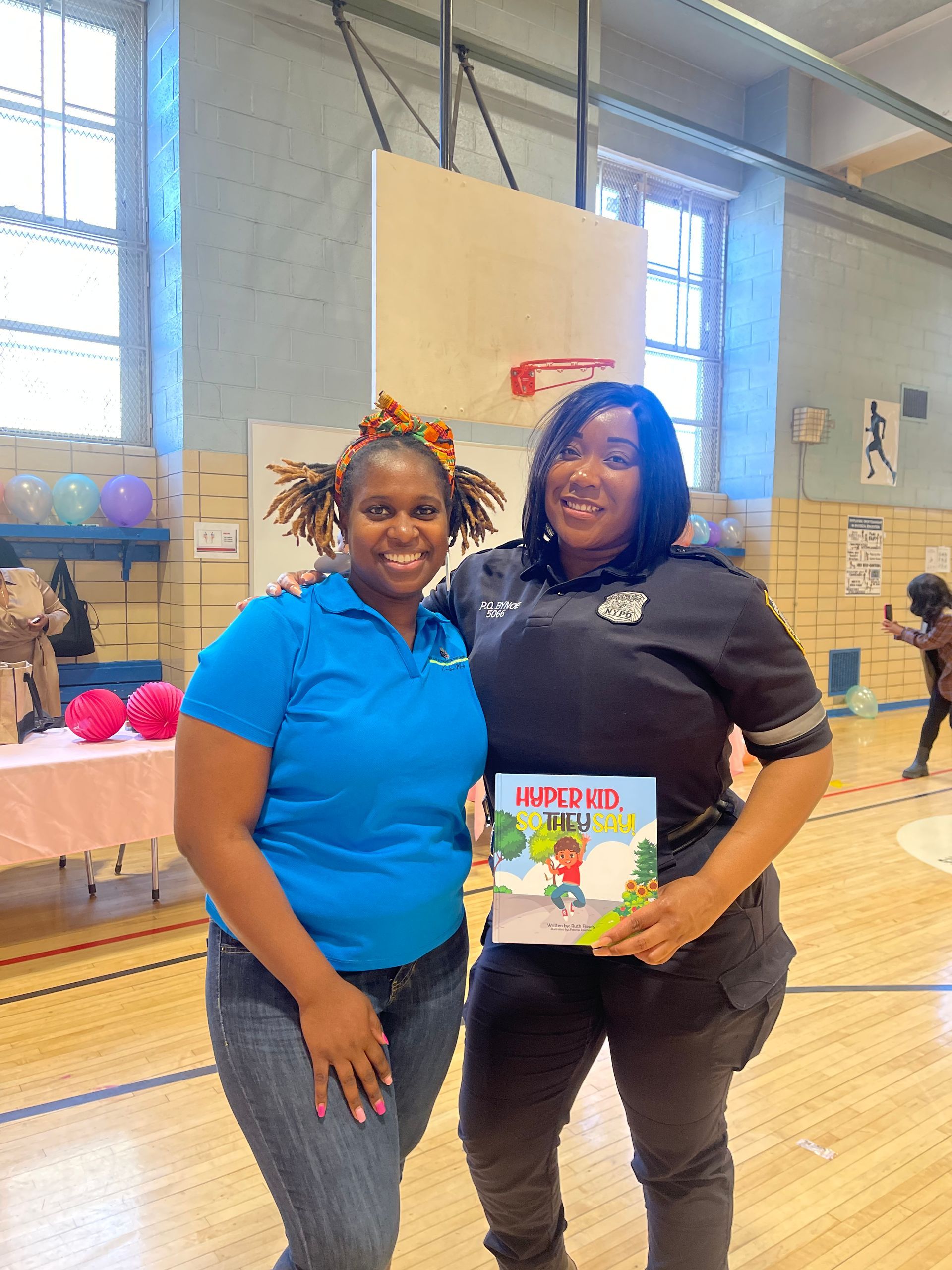 Two women pose with a children's book in an indoor gym. One wears blue; the other, black.