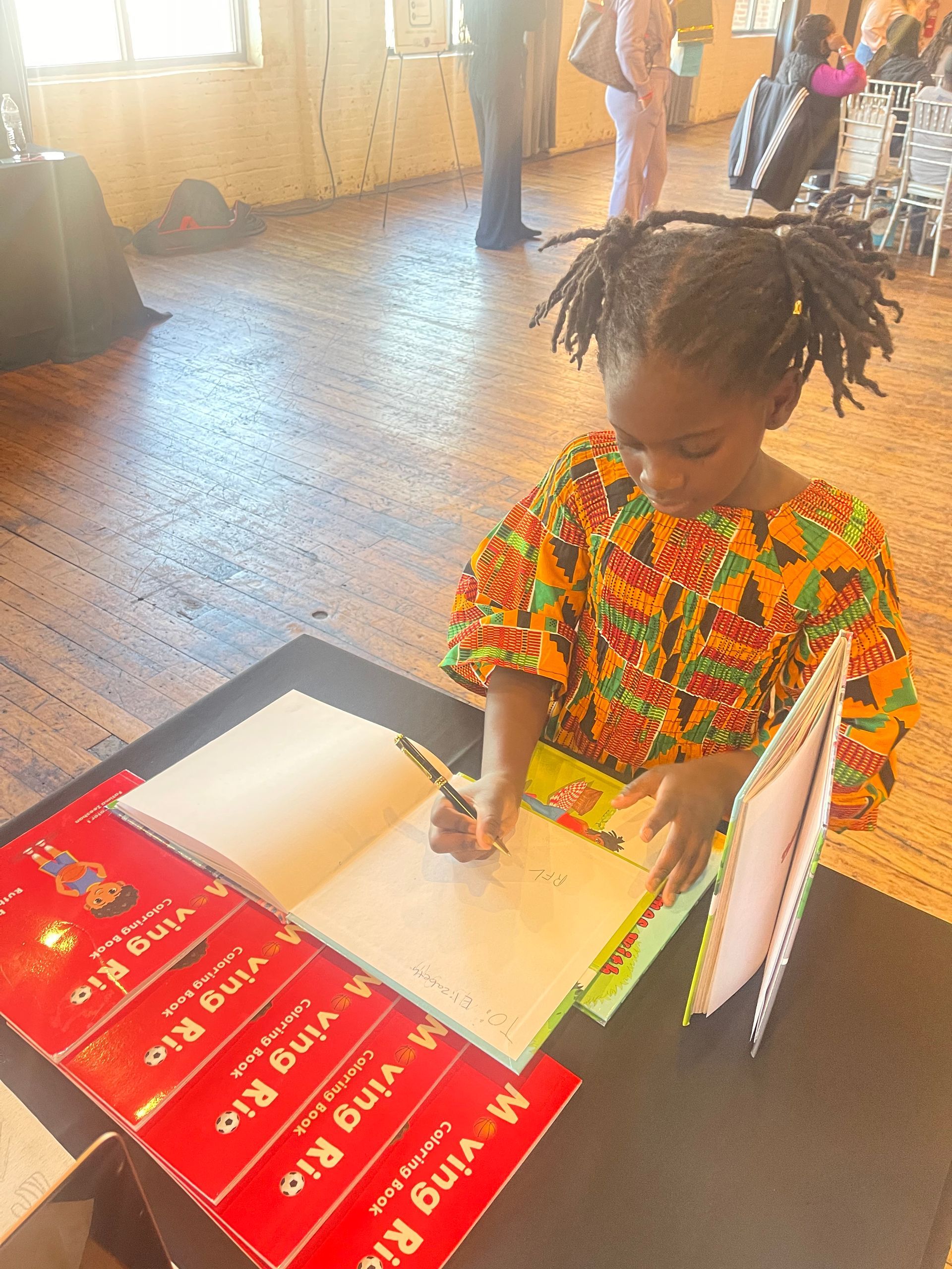 Young child in colorful patterned shirt writing in an open book. Several copies of a red book are on the table.