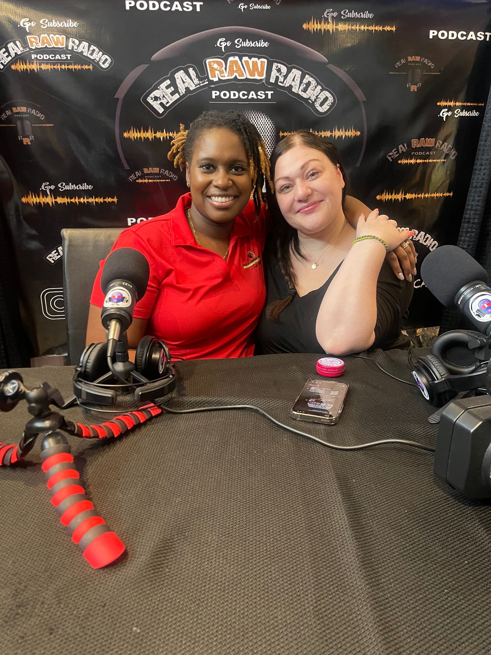 Two women smiling, seated at a podcast desk. Microphones and recording equipment are visible.
