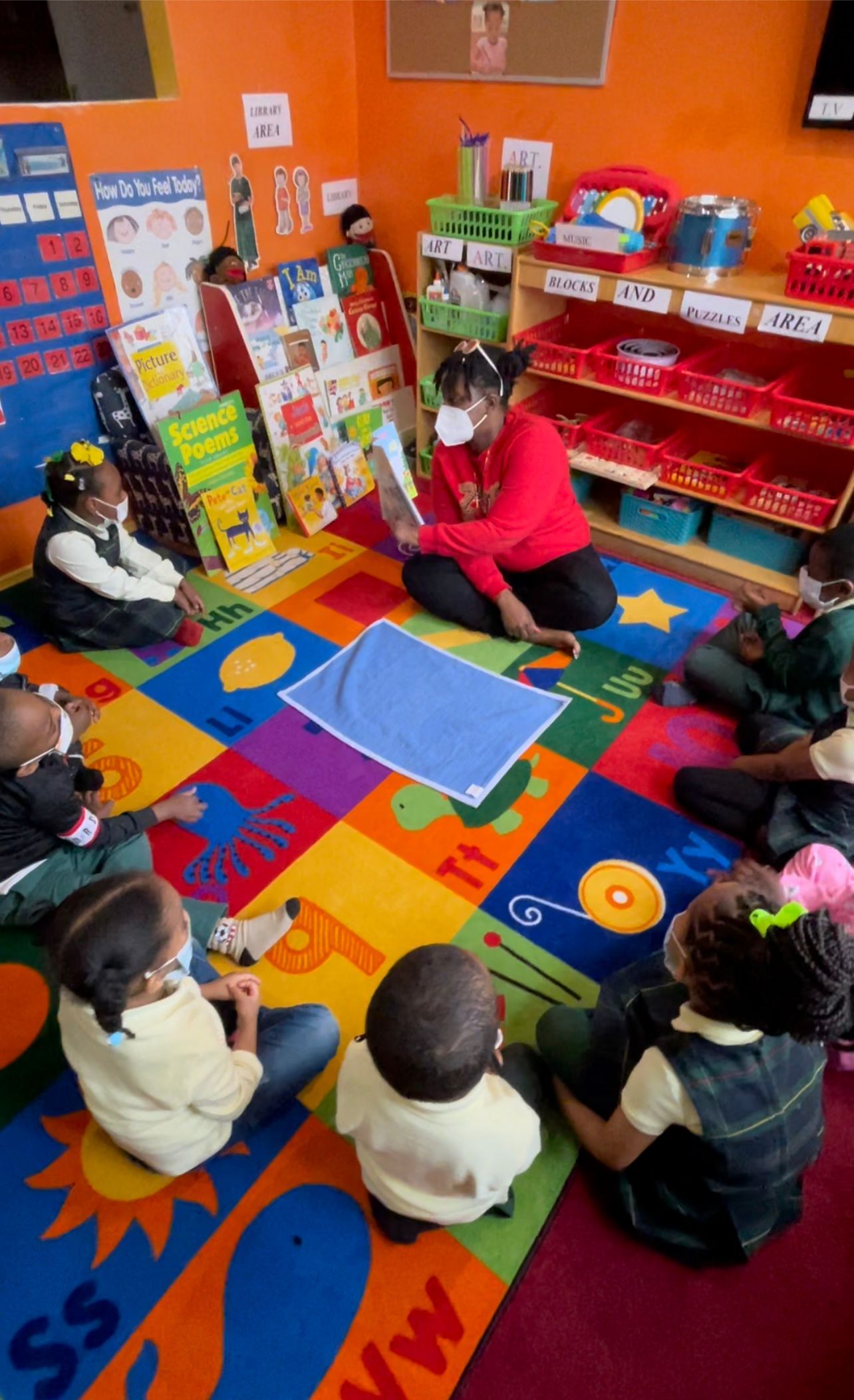 Teacher reads a book to a group of children seated on a colorful rug in a classroom.