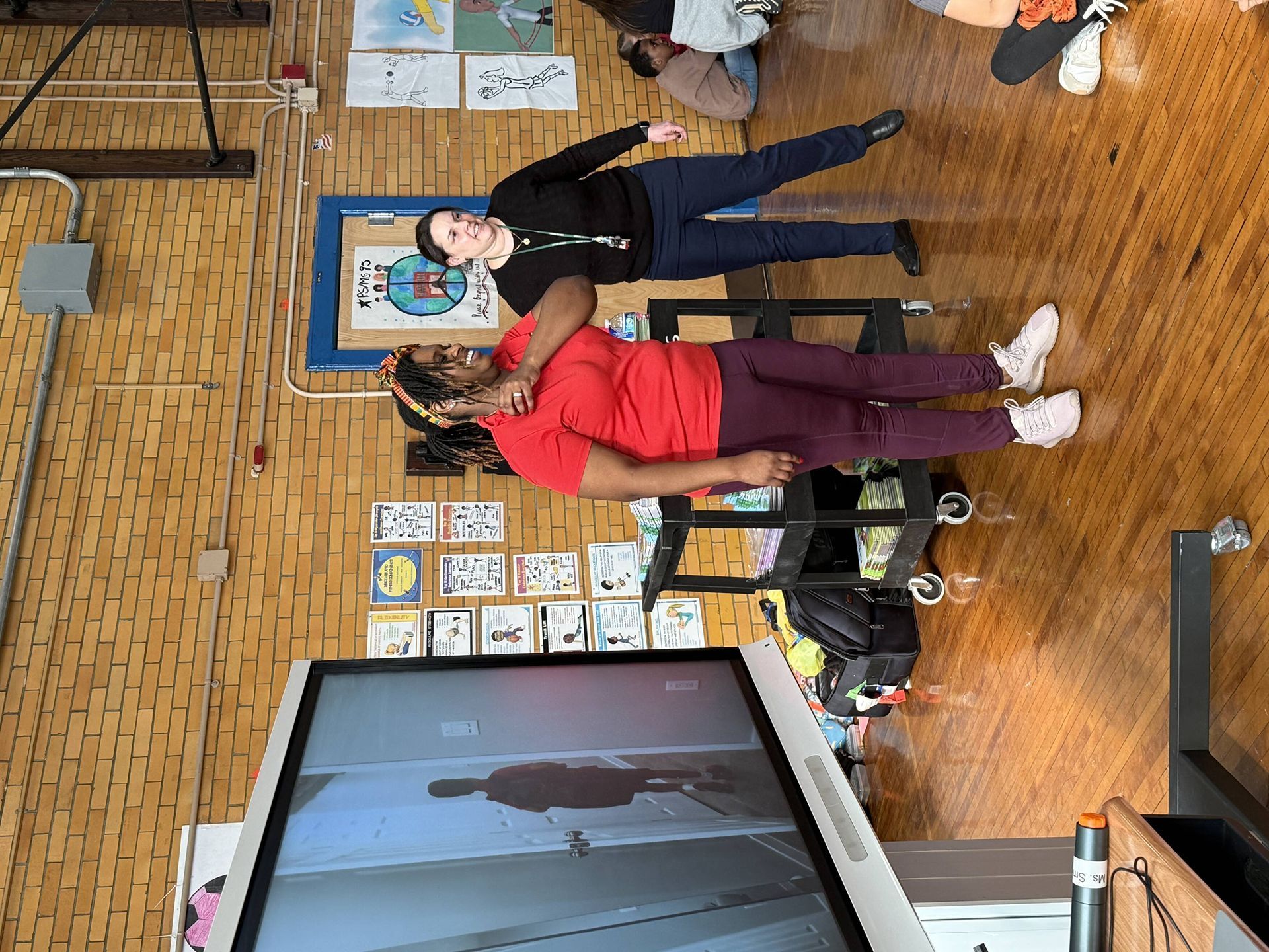 Two people practicing self-defense in a room with a large screen displaying an interior, brick wall background.