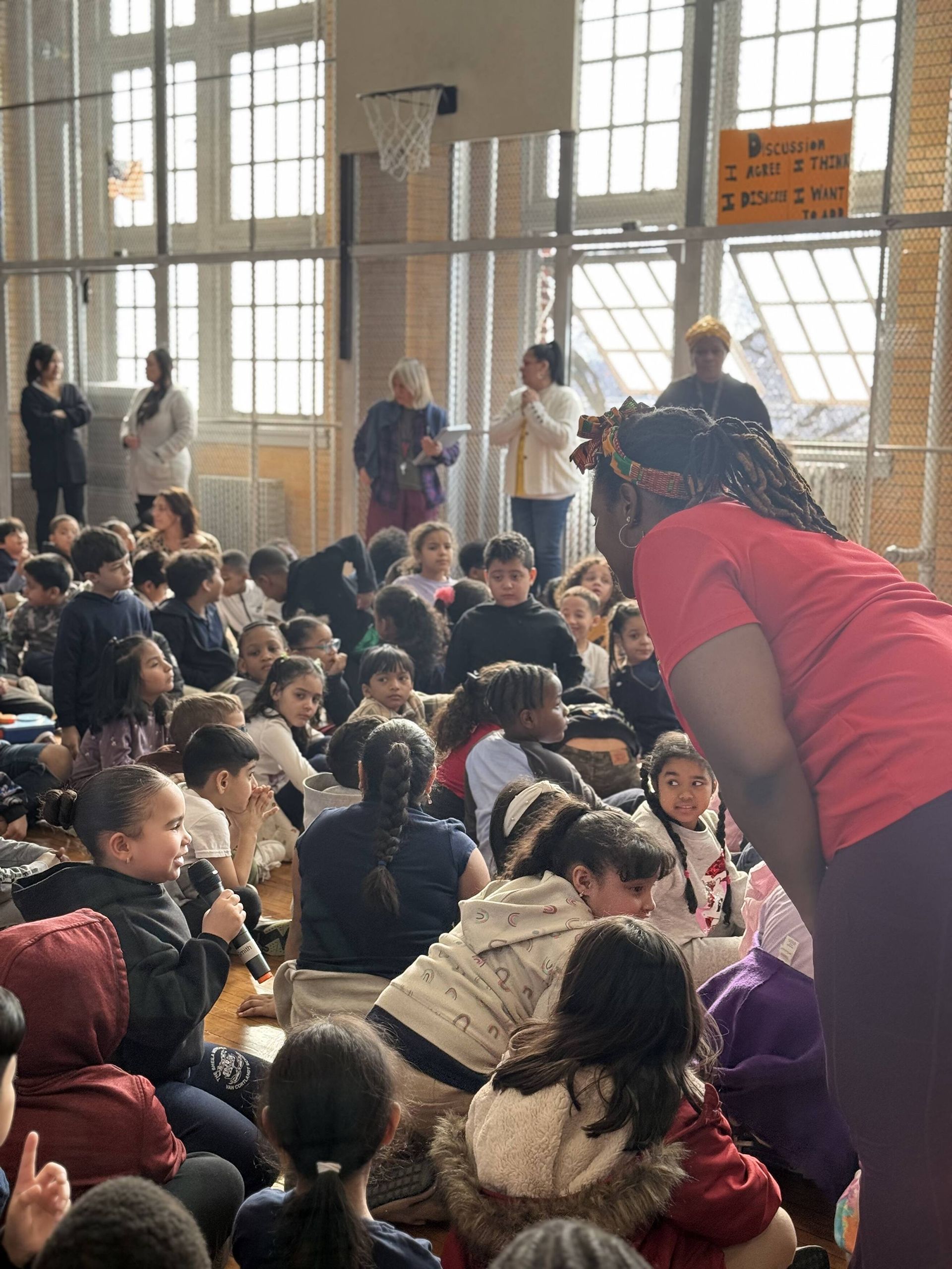 Children seated on floor, listening to a person wearing a red shirt, in a brightly lit school gymnasium.