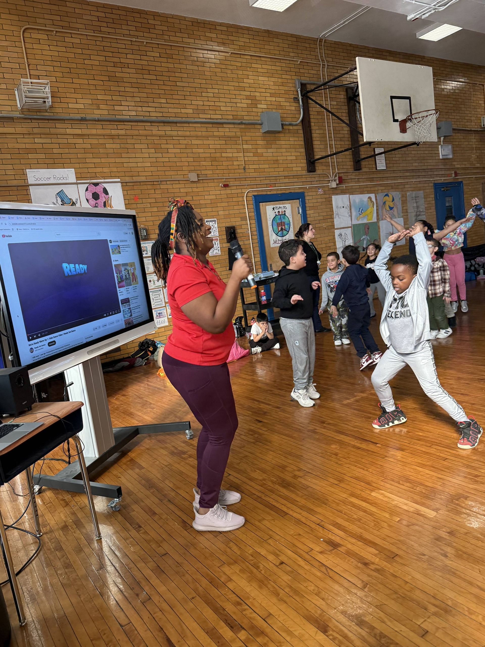 Woman in red shirt filming children exercising in a gym. Interactive whiteboard visible.
