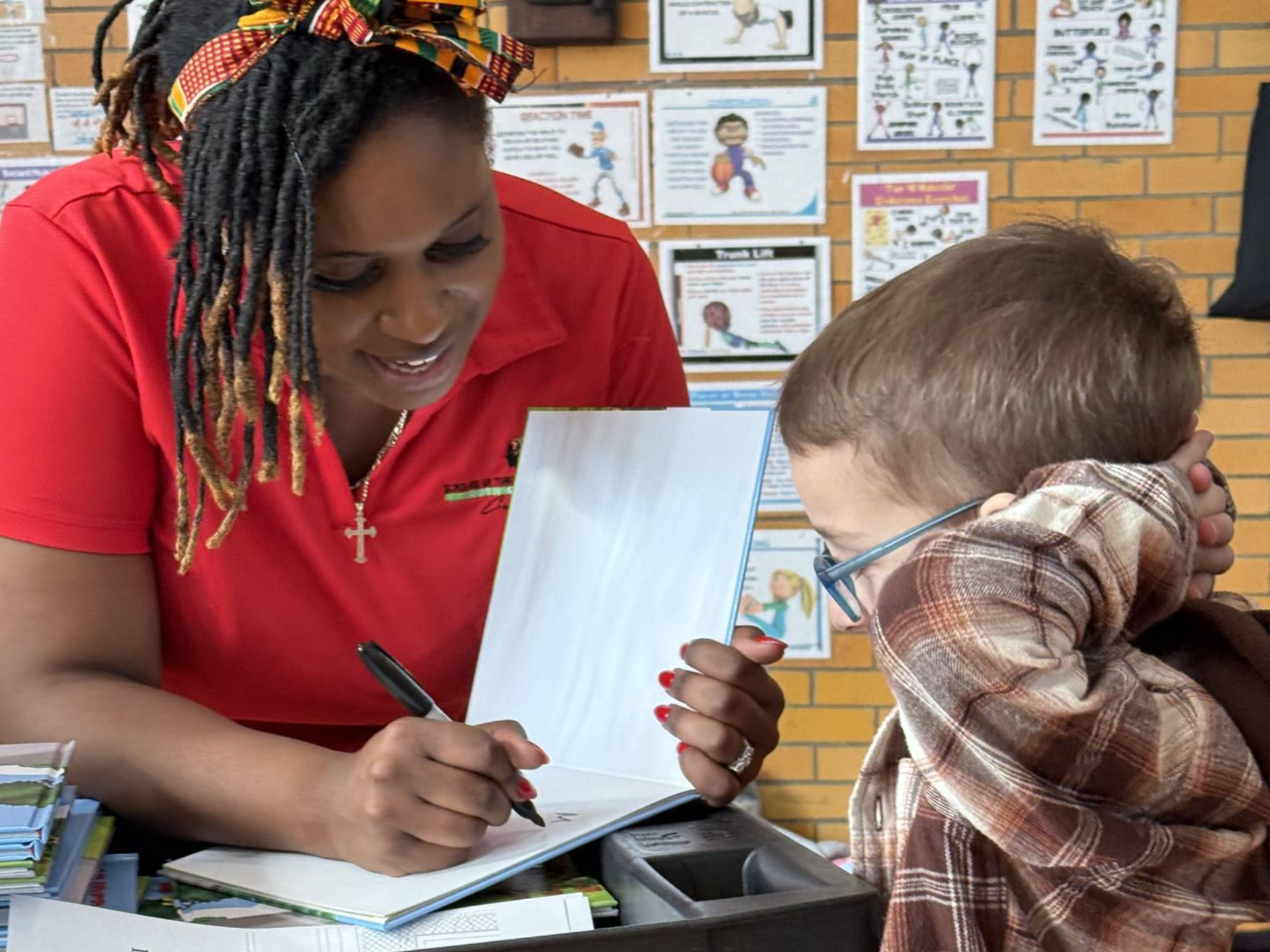 Woman in red shirt drawing in a book with a child, both seated, indoors.