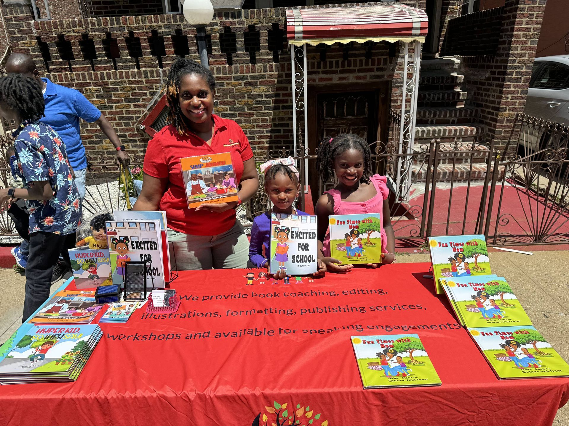 Woman and children at a book sale table, smiling, holding books. Red tablecloth and a brick building in the background.