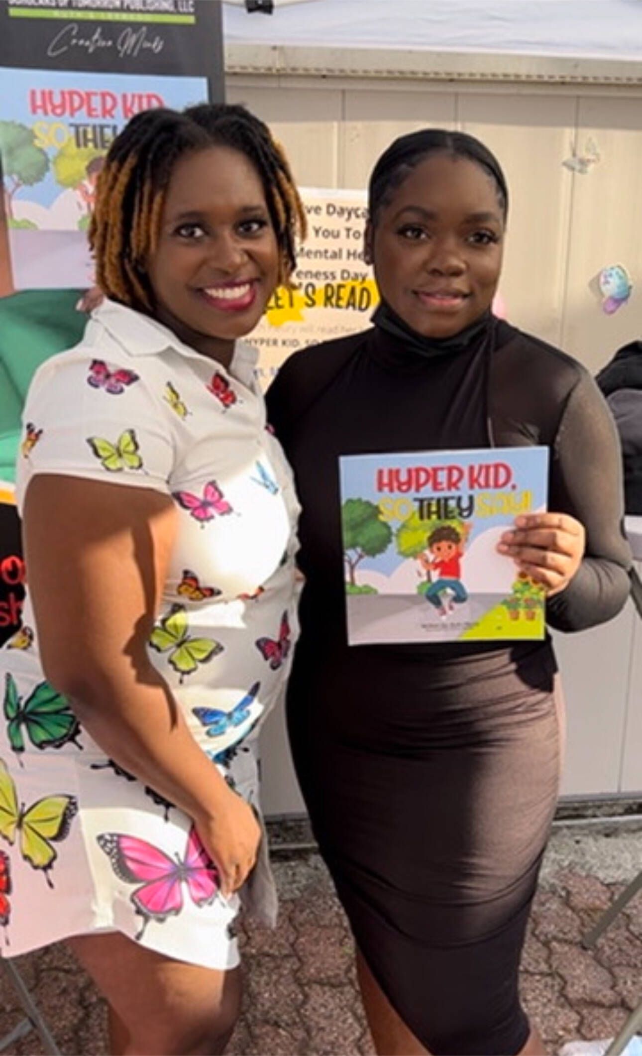 Two women pose, holding a book. One wears a butterfly print dress, the other, a dark turtleneck and skirt.